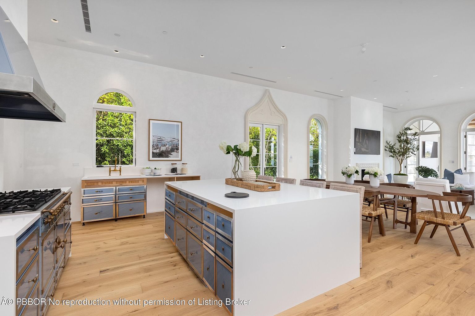 This bright and airy kitchen features a large central island with a white countertop and blue-toned cabinetry, complemented by light wood flooring throughout. The space is anchored by a professional-style range and a secondary sink area, with an open-concept layout that flows into a dining and living area. Tall arched windows and doors allow for abundant natural light, creating a sophisticated and welcoming atmosphere.