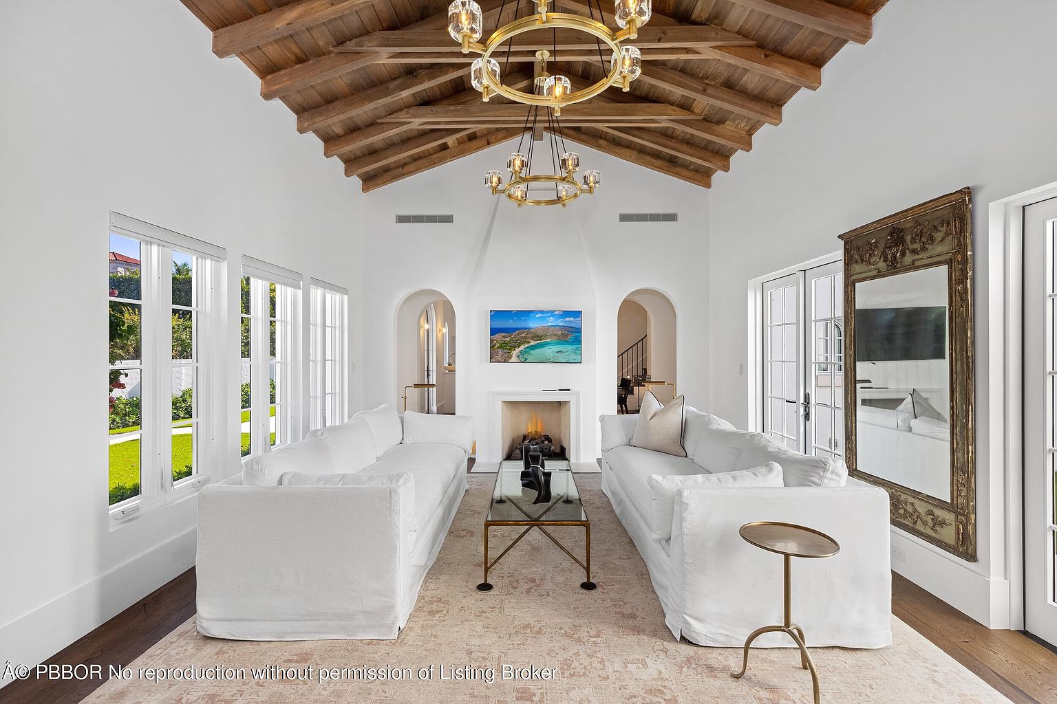 This elegant living room features a dramatic vaulted ceiling with exposed wooden beams and a central gold-toned chandelier. The space is anchored by two plush white sofas facing a central fireplace, which is set into a white wall beneath a mounted television. Large windows on the left and glass doors on the right fill the room with natural light, while a large, ornate gold-framed mirror adds a touch of sophistication to the side wall.