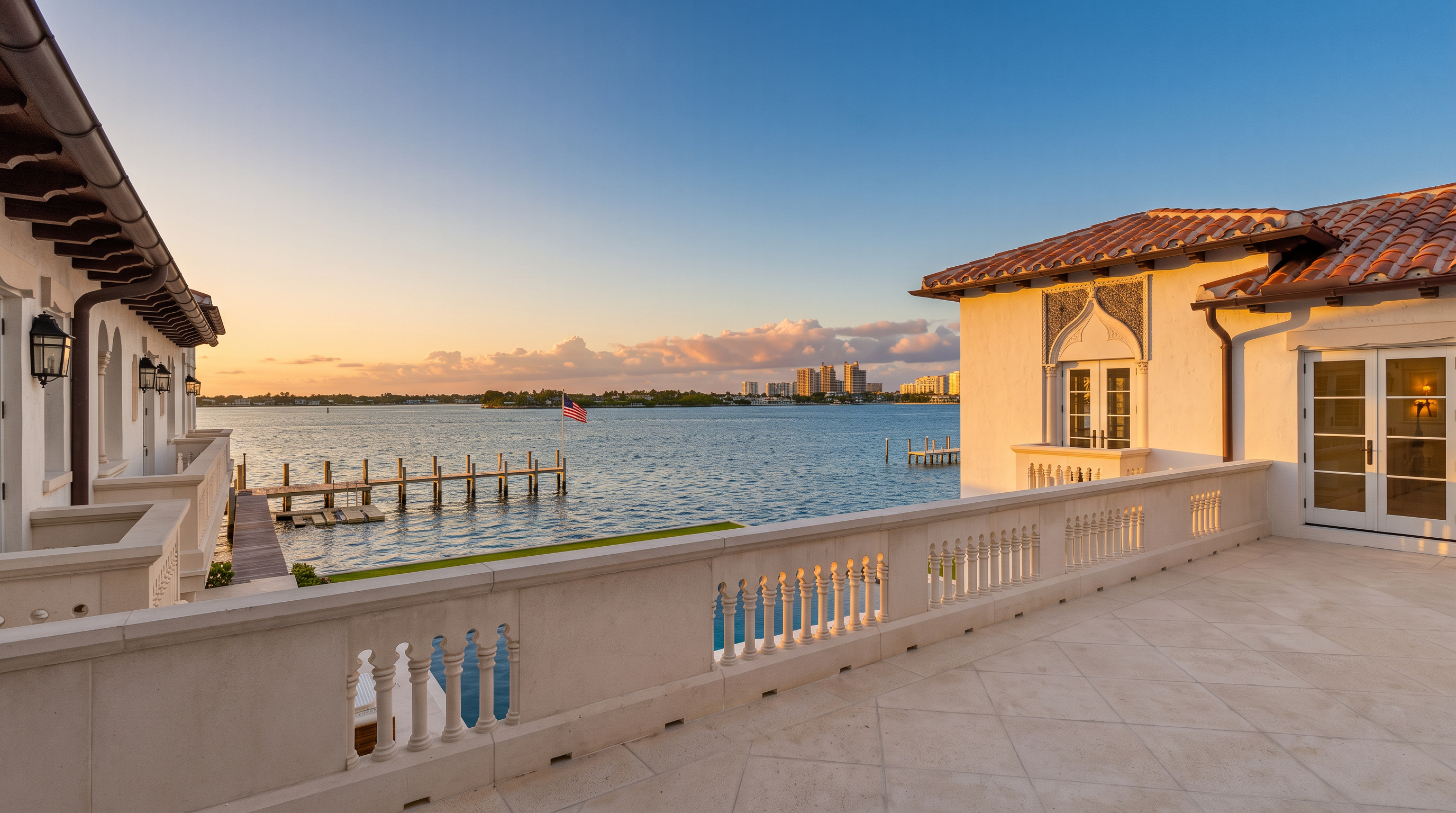 This expansive, light-colored stone balcony offers a panoramic view of a calm, blue waterway and a distant city skyline. The space is framed by classic balustrades and features elegant Mediterranean-style architecture with terracotta roof tiles and white stucco walls. A private wooden dock extends into the water, creating a serene and luxurious waterfront atmosphere.