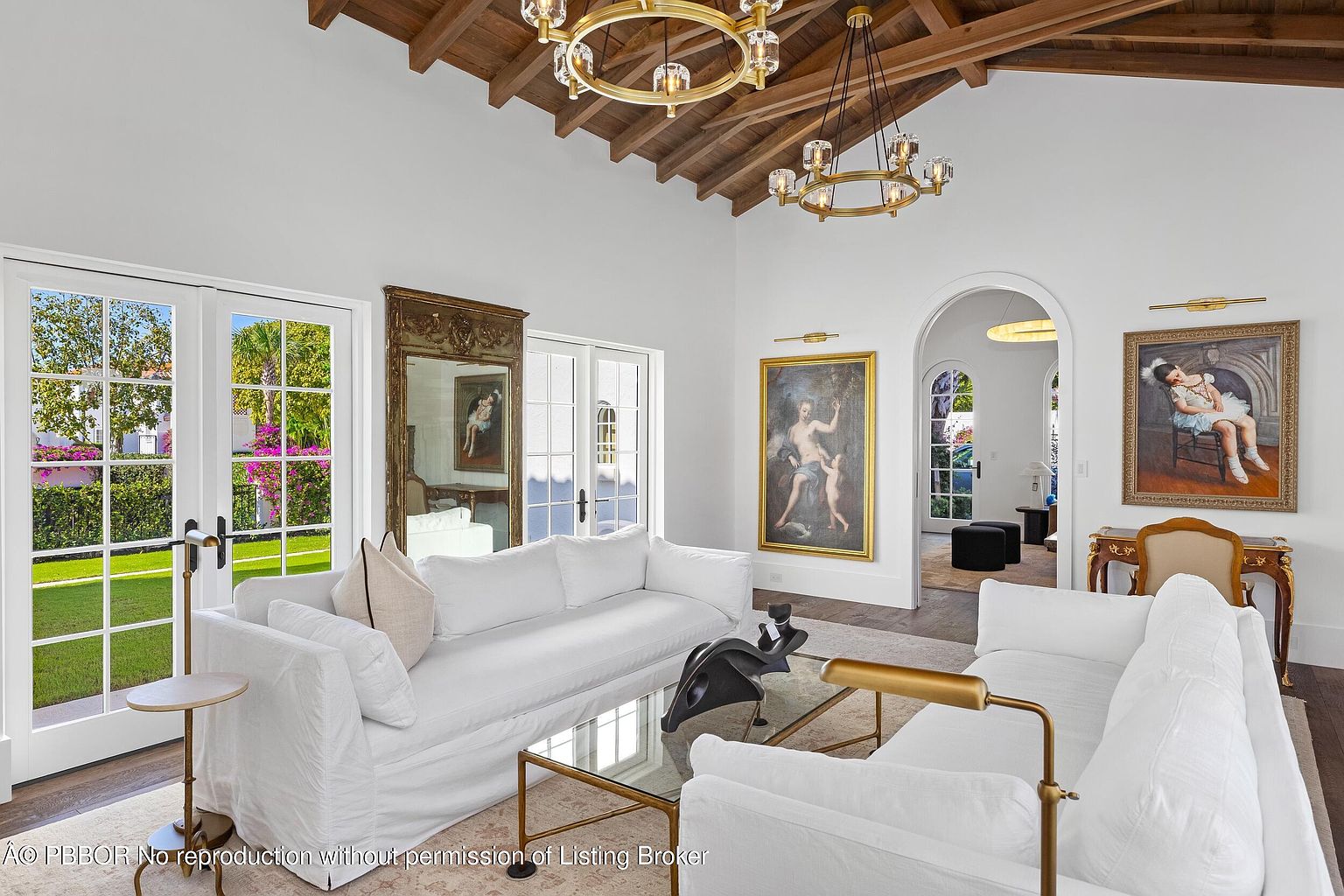 This elegant living room features a vaulted wood-beamed ceiling and two large, circular chandeliers that anchor the space. The room is furnished with two plush white sofas facing a glass-topped coffee table, complemented by classical artwork and a large ornate mirror. Tall glass doors provide abundant natural light and a view of the lush green exterior, creating a sophisticated and airy atmosphere.