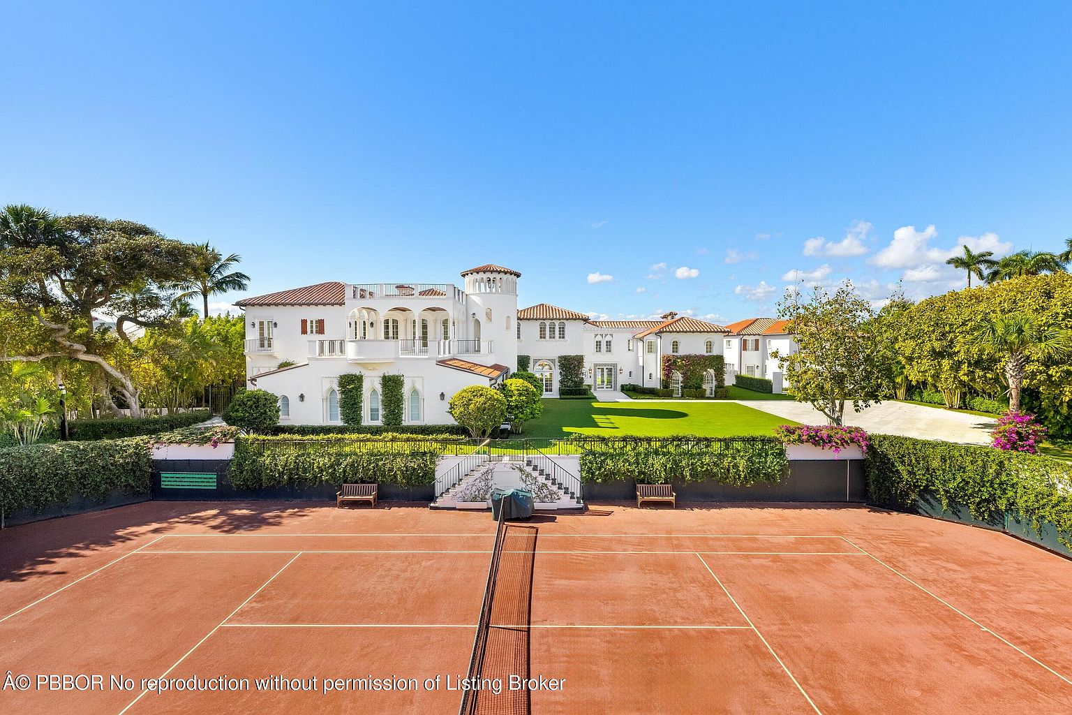 This expansive Mediterranean-style estate is captured from an elevated perspective, looking across a private clay tennis court toward the main residence. The white stucco home features multiple levels, arched architectural details, and a terracotta-tiled roof, set against a backdrop of lush green lawns and mature landscaping. The scene conveys a sense of luxury and privacy, highlighting the property's significant outdoor recreational space.