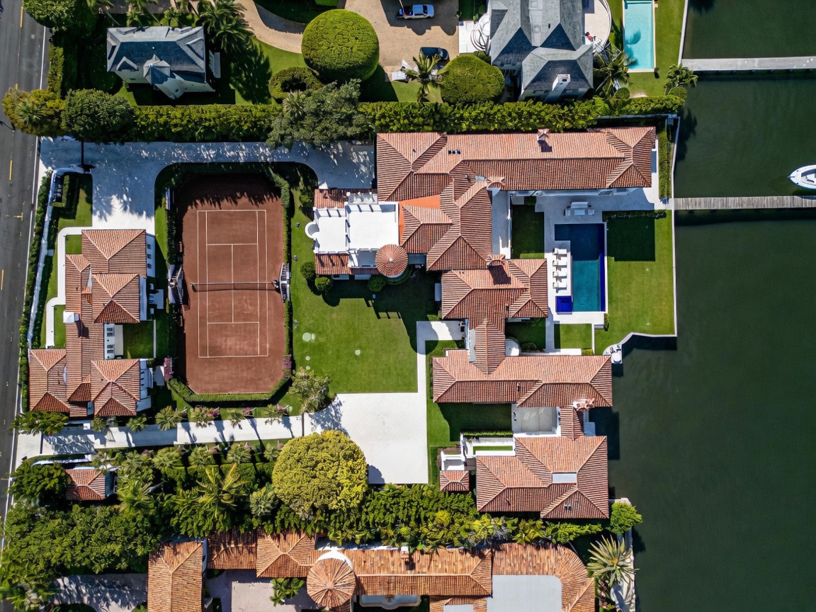 This high-angle aerial view captures a sprawling luxury waterfront estate featuring a clay tennis court, a private swimming pool, and multiple interconnected structures with terracotta-tiled roofs. The property is situated directly on a waterway, complete with a private dock and boat slip, surrounded by lush landscaping and mature trees. The layout emphasizes expansive outdoor living spaces and a sophisticated, Mediterranean-inspired architectural design.