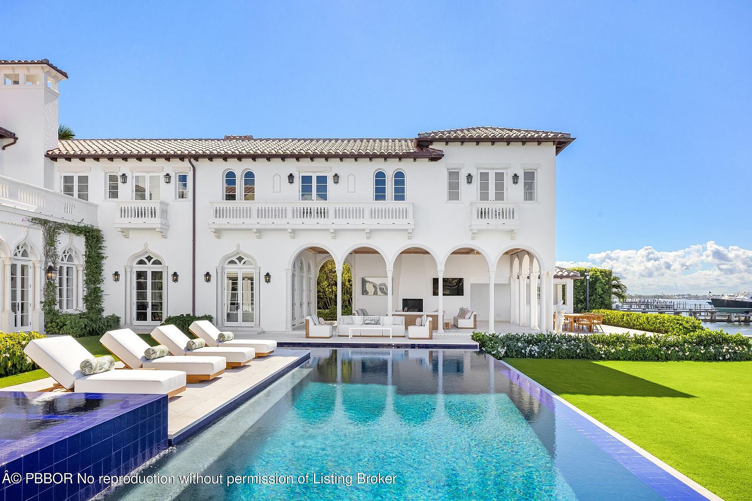 This luxurious Mediterranean-style estate features a pristine infinity-edge pool that reflects the bright blue sky and the white stucco facade of the home. The rear exterior showcases a covered loggia with comfortable outdoor seating, elegant arched architectural details, and a manicured lawn leading toward the waterfront. The perspective captures the seamless transition between the sophisticated indoor-outdoor living spaces and the serene coastal environment.
