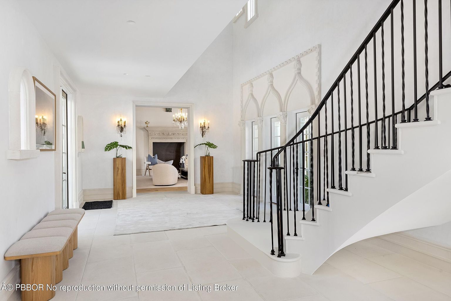 This elegant foyer features a grand staircase with a black iron railing and light-colored stone flooring. A long bench sits against the left wall, while two wooden pedestals topped with greenery frame the entrance to an adjacent living space. The bright, airy atmosphere is enhanced by white walls, decorative architectural molding, and warm wall sconces.