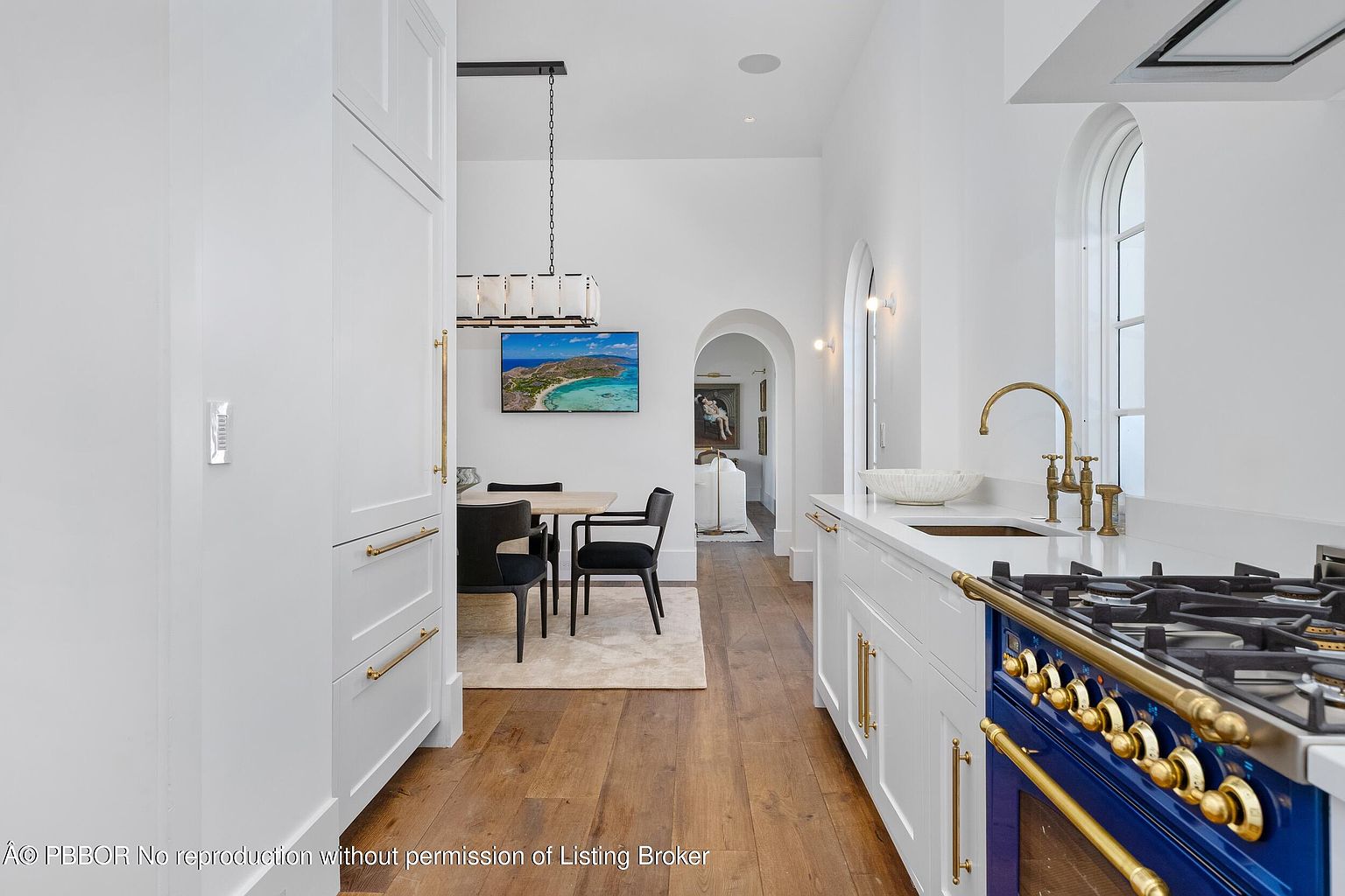 This bright, modern kitchen features crisp white cabinetry with gold hardware and a striking blue range as a focal point. The space flows seamlessly into a dining area, connected by a wide arched doorway that adds architectural interest. Warm wood flooring runs throughout, creating a cohesive and inviting atmosphere.