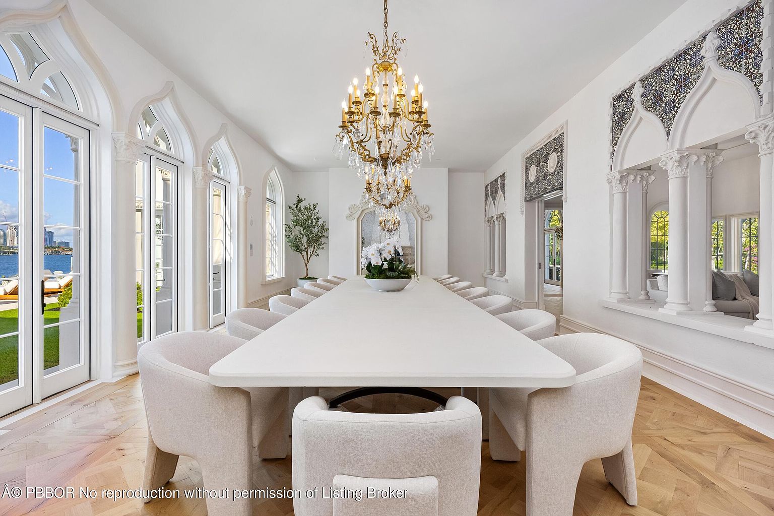This elegant dining room features a long, minimalist white table surrounded by upholstered chairs, centered beneath an ornate gold chandelier. The space is defined by its bright, white walls, intricate arched window frames, and light-toned herringbone wood flooring. A view through the tall windows reveals a waterfront setting, while decorative tile accents above the arched wall niches add a touch of architectural detail.