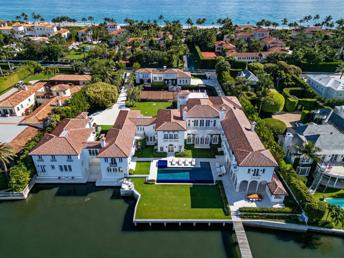 This high-angle aerial view captures a sprawling Mediterranean-style estate situated directly on a waterfront canal. The property features a large swimming pool, a manicured lawn, a private dock, and a tennis court, all surrounded by lush tropical landscaping. The architecture is defined by its white stucco walls and classic terracotta tile roofs, presenting a luxurious and expansive coastal residence.
