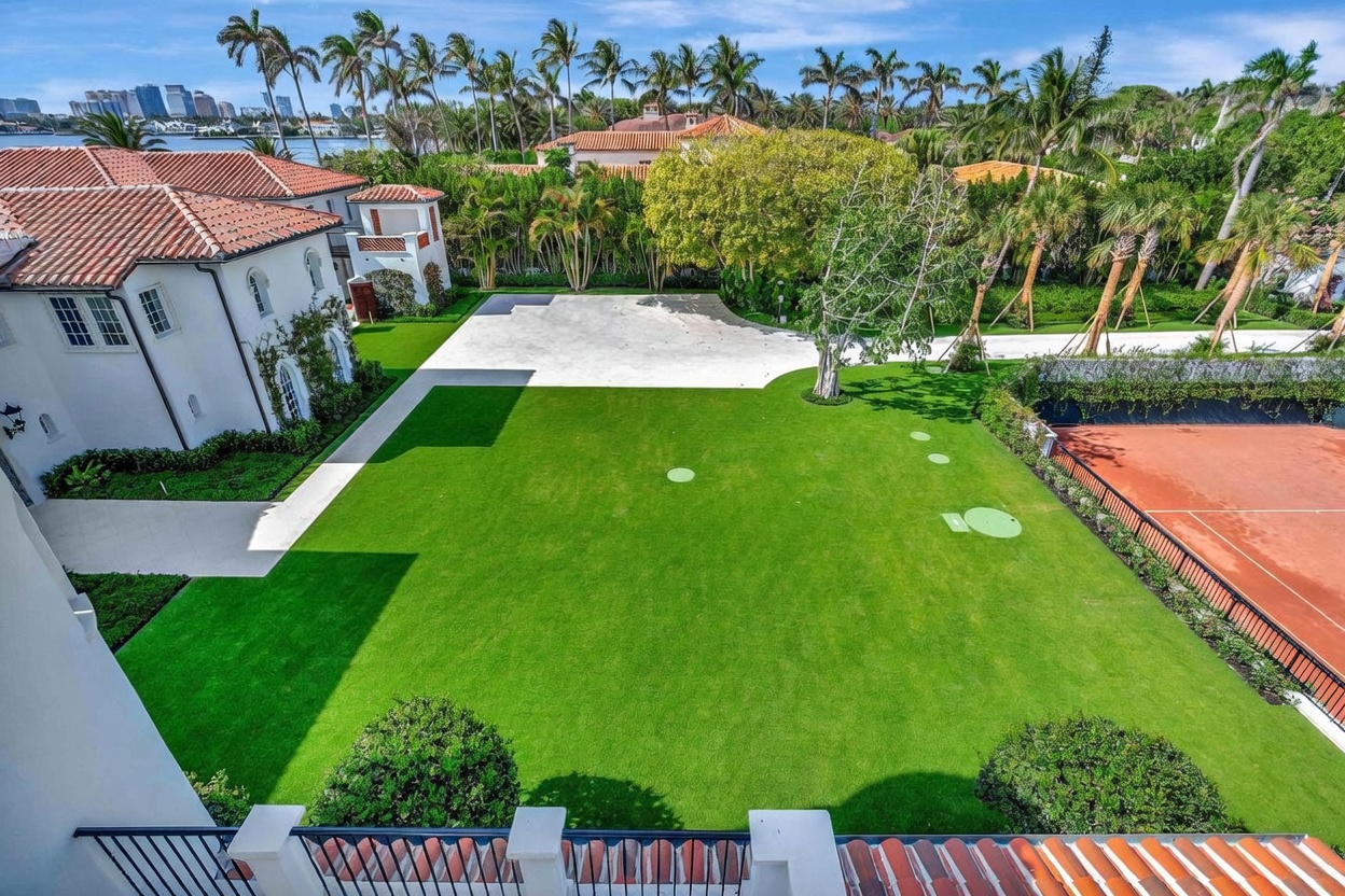 This high-angle aerial view captures a sprawling, meticulously landscaped estate featuring a large, manicured lawn with putting green elements. To the right, a clay tennis court is bordered by lush greenery, while a paved driveway area sits adjacent to the main residence, which showcases classic Mediterranean-style architecture with a terracotta tile roof. The background reveals a scenic waterfront setting with distant city buildings, emphasizing the property's prime location and expansive outdoor living space.