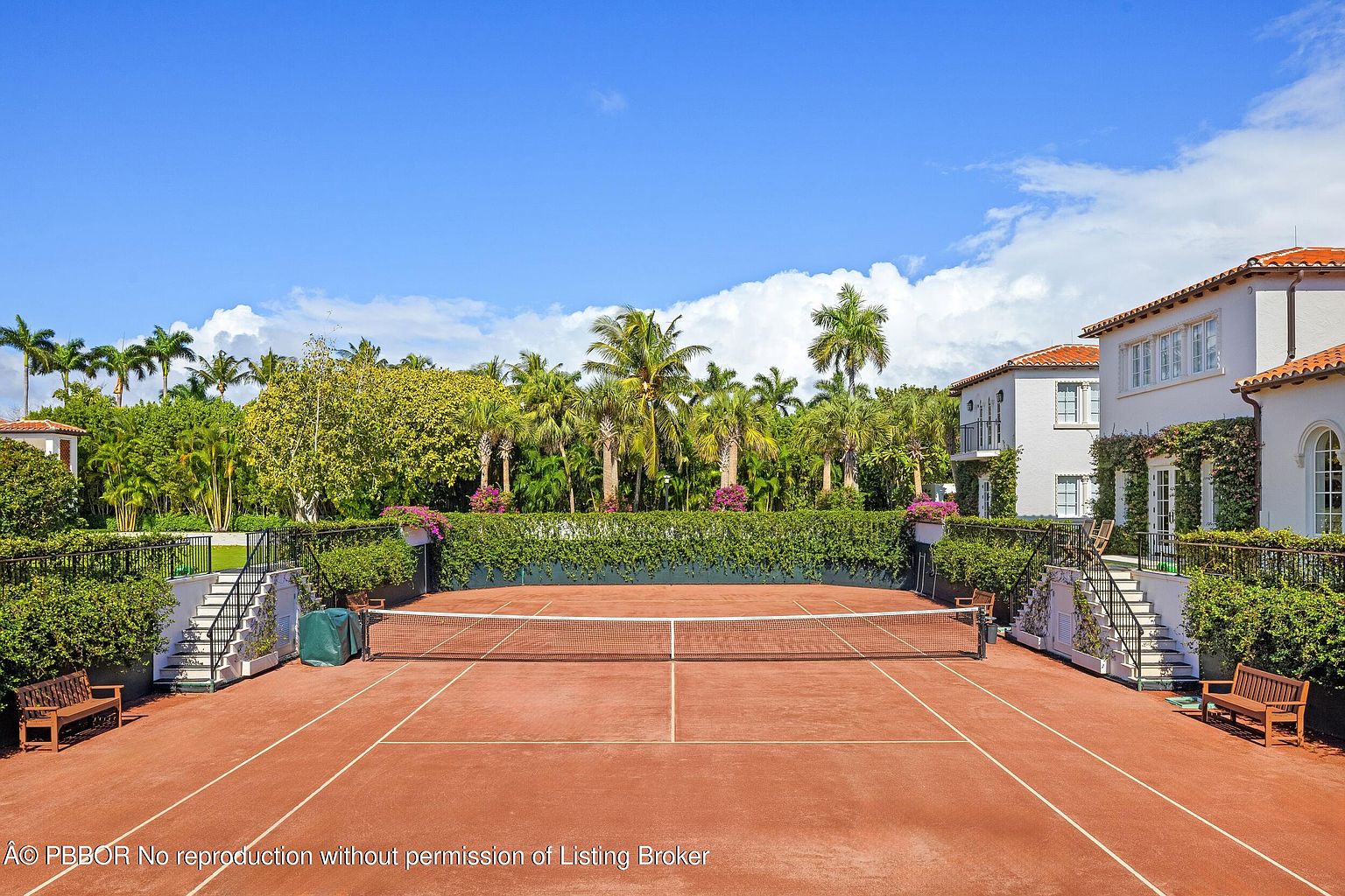 This image showcases a private clay tennis court nestled within a lush, tropical landscape. The court is framed by manicured hedges, mature palm trees, and a grand Mediterranean-style residence with white stucco walls and a terracotta tile roof. The perspective is elevated, providing a clear view of the court's layout, the surrounding greenery, and the architectural details of the adjacent home.