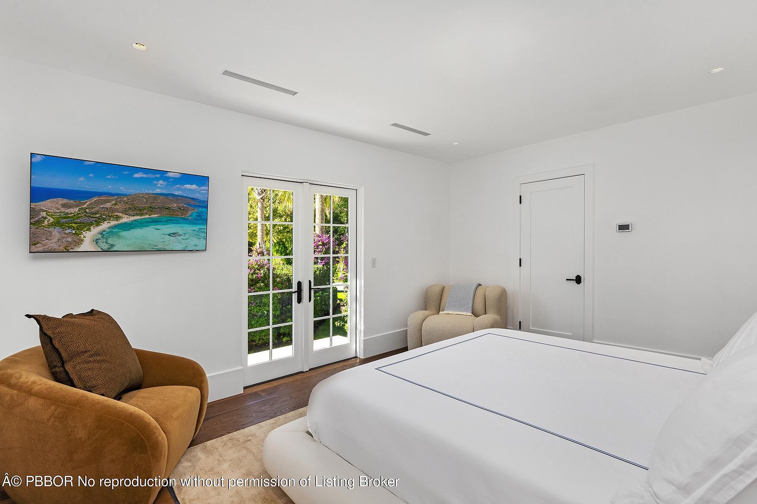 This bright and airy primary bedroom features crisp white walls, dark wood flooring, and a plush bed with white linens. A large television is mounted on the wall, while a pair of French doors provides access to a lush, green outdoor space. The room is furnished with a comfortable, warm-toned armchair in the foreground and a secondary neutral-toned chair near the corner, creating a serene and inviting atmosphere.