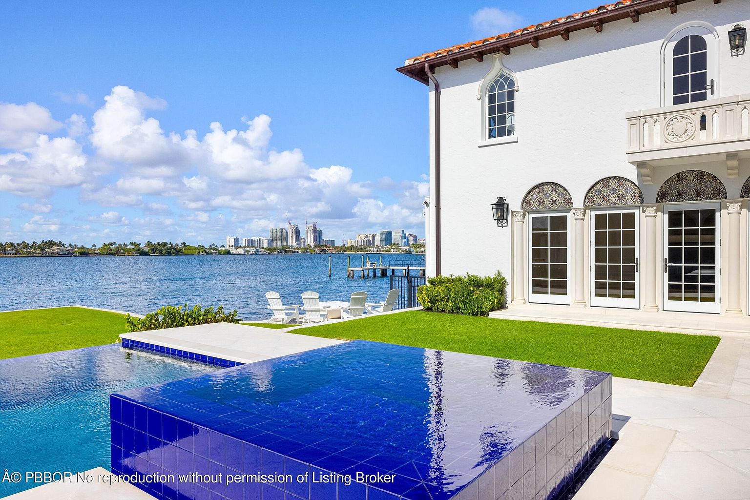 This luxurious waterfront property features a vibrant blue tiled infinity-edge pool that seamlessly blends with the expansive bay view. The white Mediterranean-style home includes arched glass doors and decorative tile accents, opening onto a manicured green lawn with white lounge chairs. The scene captures a serene, high-end coastal lifestyle with a clear blue sky and a distant city skyline in the background.