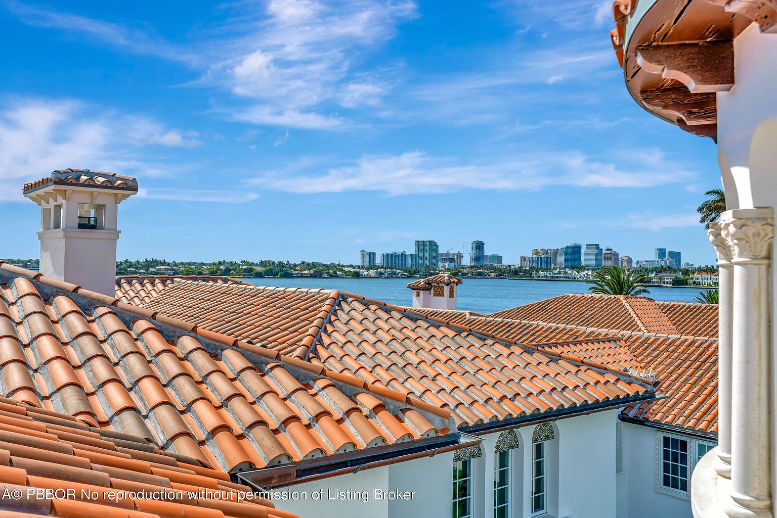 This elevated perspective captures a scenic view from a balcony overlooking a Mediterranean-style home with terracotta-tiled roofs. The foreground features the textured orange roof tiles and a decorative chimney, while the background showcases a tranquil bay with a distant city skyline under a bright blue sky. The architectural style emphasizes classic coastal elegance, providing a serene and expansive outlook.