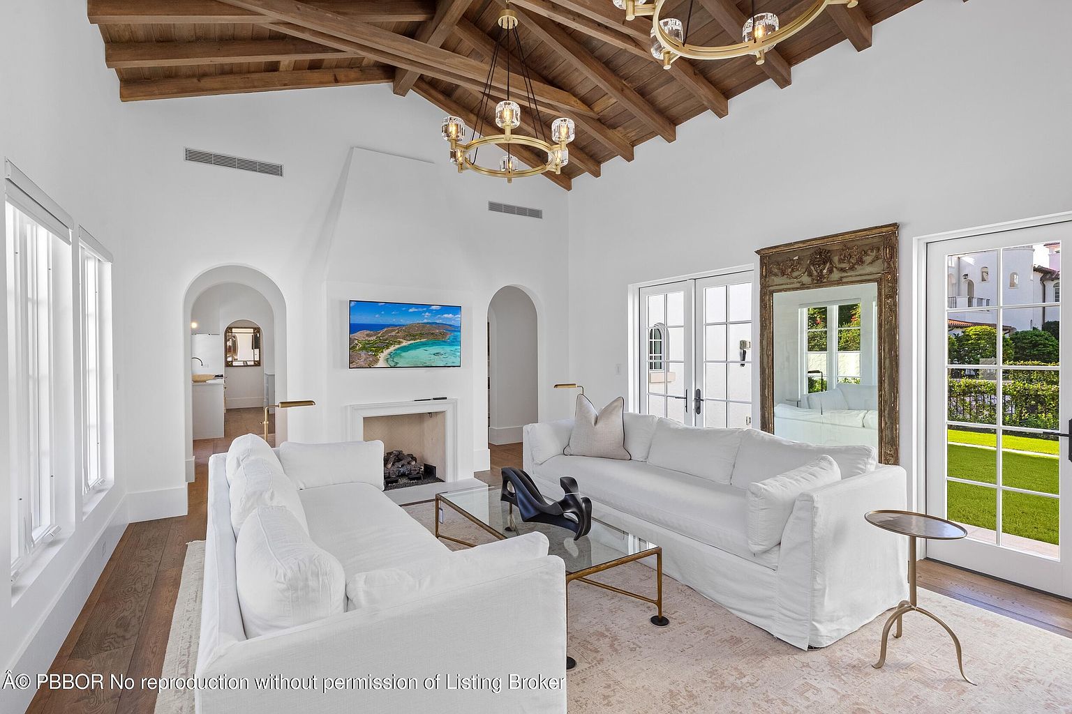 This bright and airy living room features a vaulted wood-beamed ceiling and a central fireplace with a tall, white surround. Two white sofas face each other over a glass-topped coffee table, creating a symmetrical and inviting seating area. Large glass doors and windows provide ample natural light and views of the outdoor greenery, while a large ornate mirror adds a touch of elegance to the space.