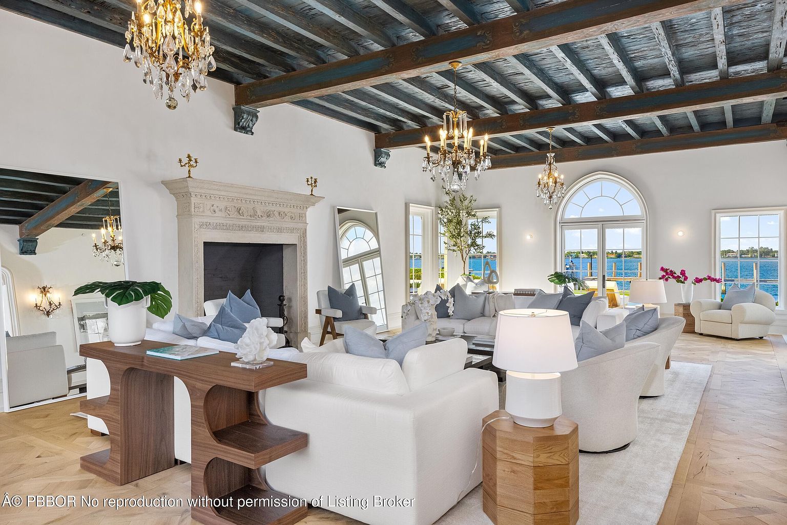 This expansive living room features a sophisticated design with a dark, exposed beam ceiling and light-toned herringbone wood flooring. A large stone fireplace serves as a central focal point, complemented by plush white seating arrangements and elegant crystal chandeliers. The space is bathed in natural light from large arched windows and glass doors that provide a clear view of the waterfront beyond.
