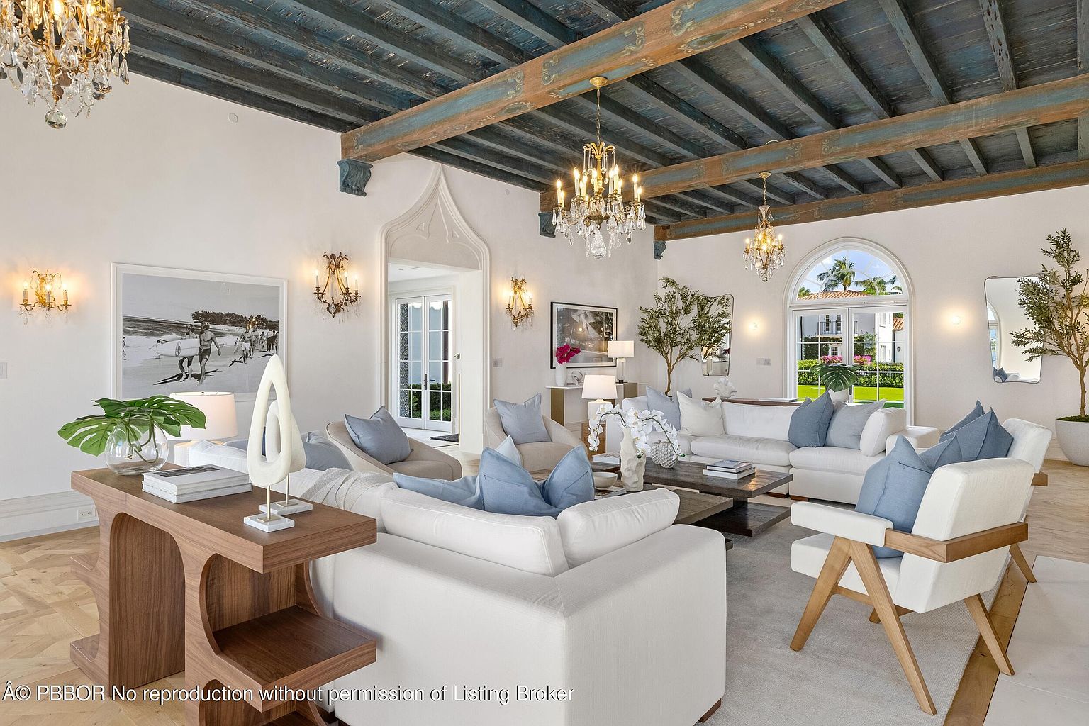This elegant living room features a striking blue-painted exposed beam ceiling and a light-toned herringbone wood floor. The space is furnished with plush white sofas accented by soft blue pillows, centered around a low wooden coffee table. Large arched glass doors provide a view of the outdoor greenery, while multiple crystal chandeliers add a touch of luxury to the bright, airy atmosphere.