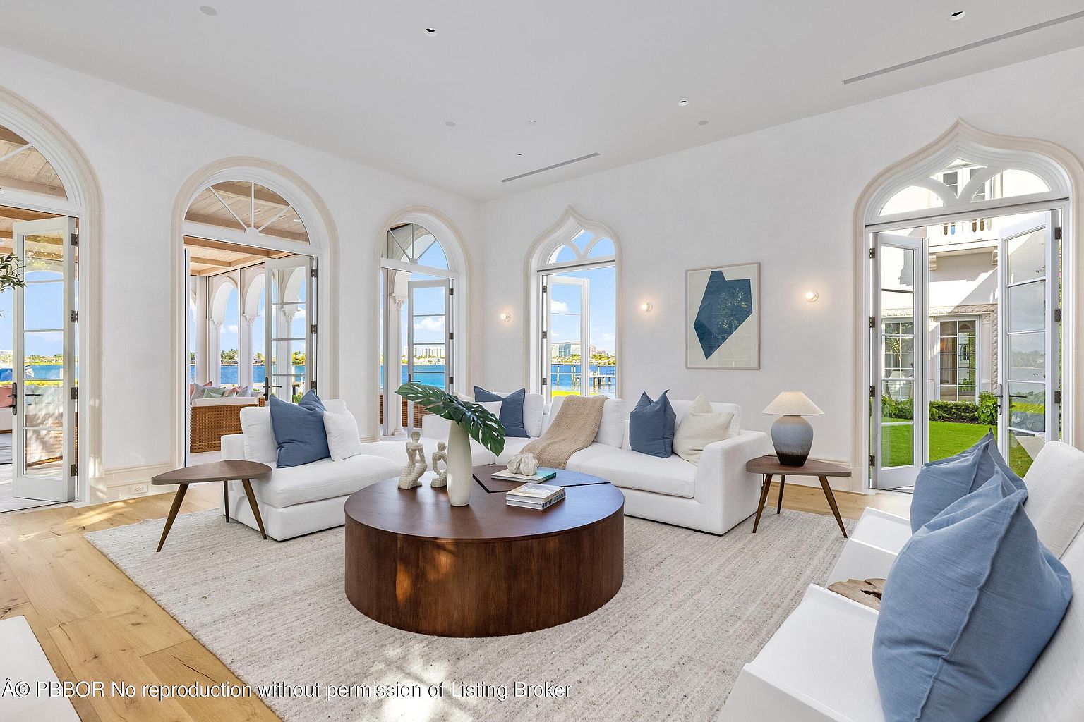 This bright and airy living room features a neutral color palette with plush white sofas accented by blue throw pillows. A large, round wooden coffee table serves as the centerpiece, while multiple arched glass doors provide abundant natural light and seamless access to the outdoor patio and water views. The space is defined by its elegant architectural arches, light wood flooring, and a clean, sophisticated aesthetic.