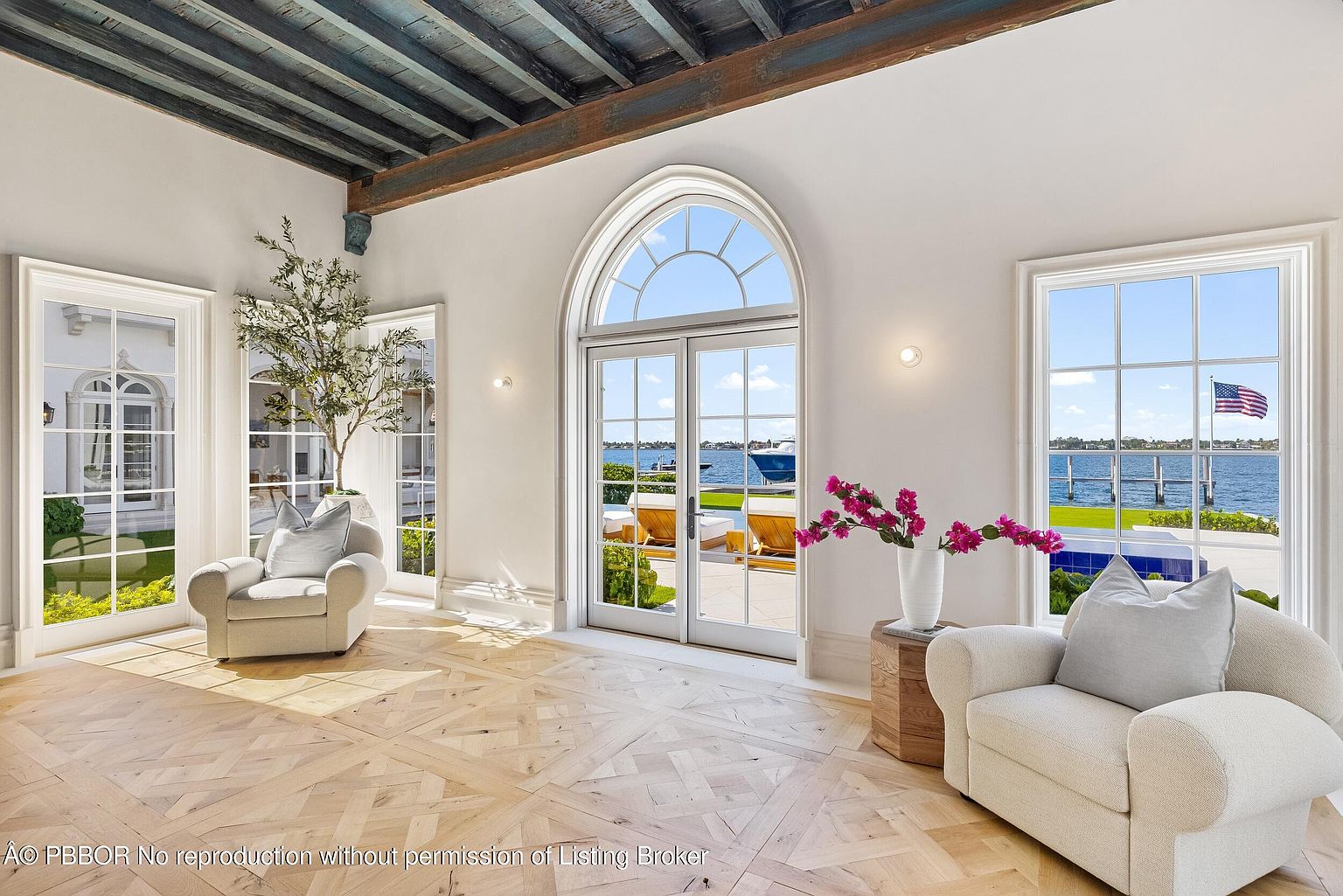 This bright and airy living space features elegant herringbone-patterned wood flooring and a dramatic exposed beam ceiling. Two plush, cream-colored armchairs are positioned to enjoy the natural light streaming through large, arched glass doors that frame a serene waterfront view. The room is accented by a potted olive tree and a vase of vibrant pink flowers, creating a sophisticated and tranquil atmosphere.