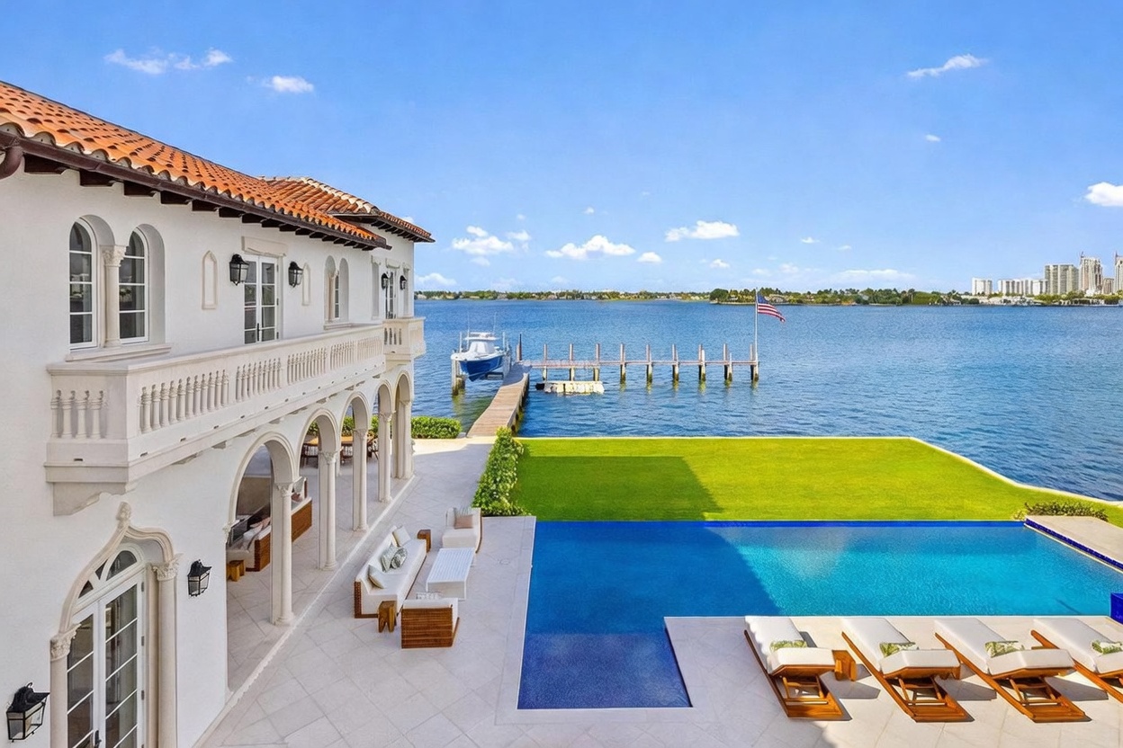 This stunning Mediterranean-style estate features a sprawling rear terrace overlooking a private infinity-edge pool and the expansive waterfront beyond. The home showcases elegant arched architectural details, a covered loggia with comfortable outdoor seating, and a manicured lawn that transitions seamlessly into the blue water. A private dock with a boat extends into the bay, creating a luxurious and serene coastal living environment.