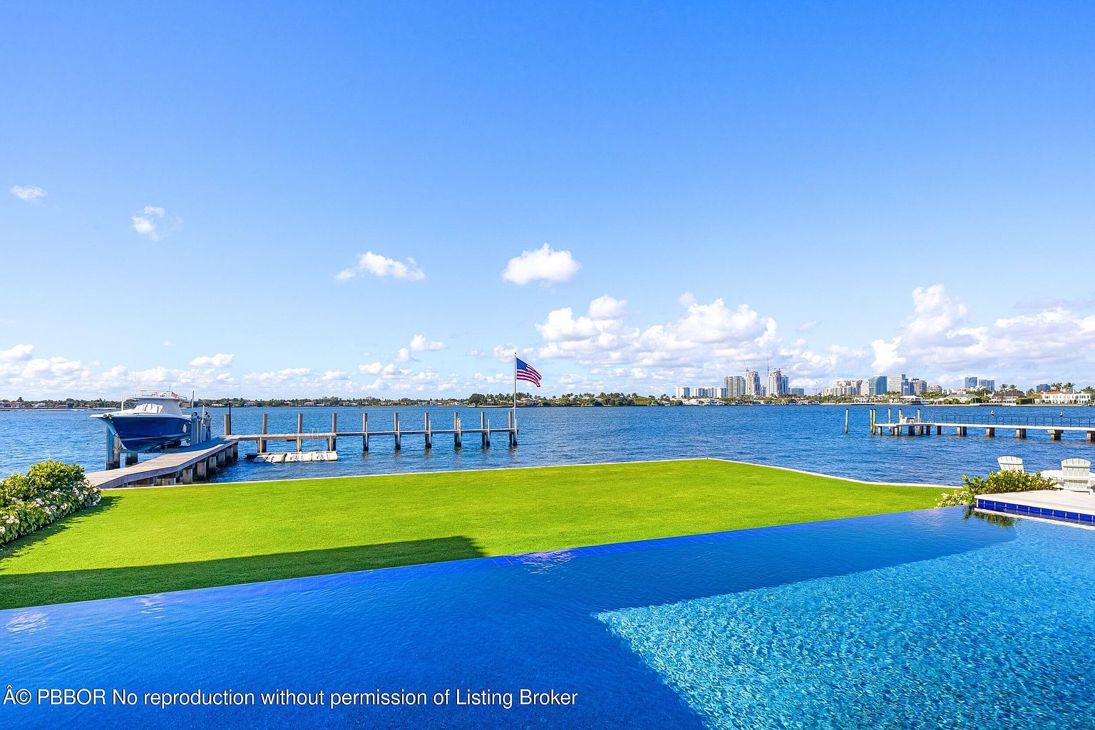 This stunning waterfront property features a luxurious infinity-edge pool that seamlessly blends into the expansive blue bay beyond. A manicured green lawn separates the pool deck from a private wooden dock, where a boat is moored under a clear, sunny sky. In the distance, a city skyline is visible across the water, creating a sophisticated and serene coastal atmosphere.