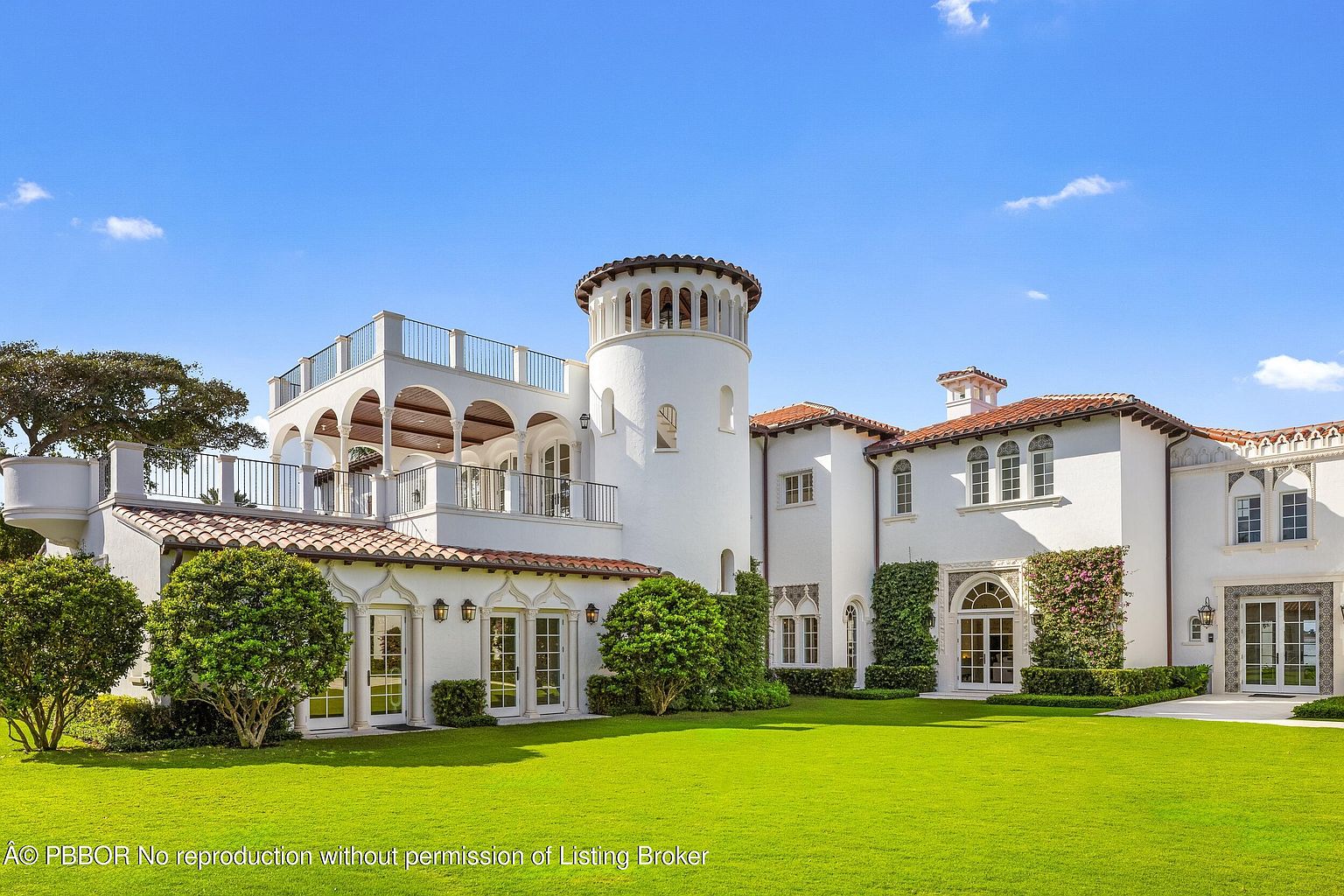 This Mediterranean-style estate features a striking white stucco facade with a prominent central tower and terracotta roof tiles. The rear elevation showcases multiple levels of balconies and terraces, complemented by lush green landscaping and manicured lawns. The architecture incorporates elegant arched windows and doorways, creating a sophisticated and expansive outdoor living space.