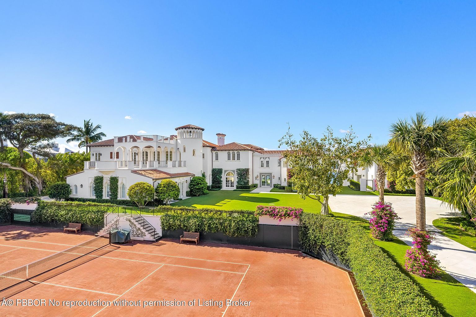 This expansive Mediterranean-style estate is captured from an elevated perspective, showcasing a meticulously manicured lawn and a private clay tennis court in the foreground. The white stucco residence features a classic red-tile roof, multiple arched balconies, and a distinctive corner tower, exuding an air of luxury and architectural elegance. The scene is framed by lush greenery, vibrant bougainvillea, and a paved driveway, creating a serene and high-end residential atmosphere.