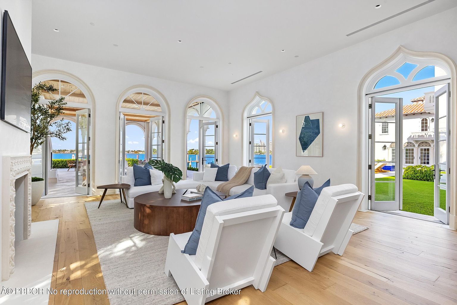 This bright and airy living room features a neutral color palette with light wood flooring and crisp white walls. The space is furnished with a large sectional sofa and two modern white armchairs, centered around a circular wood coffee table. Multiple arched glass doors provide abundant natural light and seamless access to the outdoor patio and waterfront views.