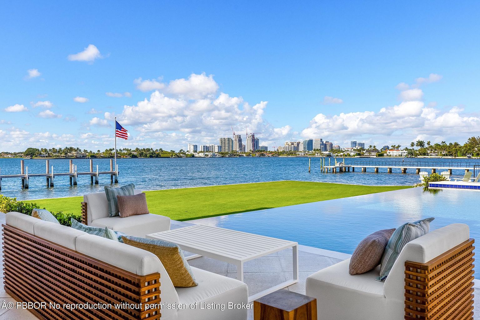 This luxurious outdoor living space features a sleek infinity-edge pool that seamlessly blends into the expansive waterfront view. The patio is furnished with modern, comfortable seating arrangements, including a white sofa with plush cushions, perfect for enjoying the panoramic vista of the bay and distant city skyline. A manicured green lawn separates the pool deck from the water, where a private dock extends into the calm bay under a bright, clear sky.