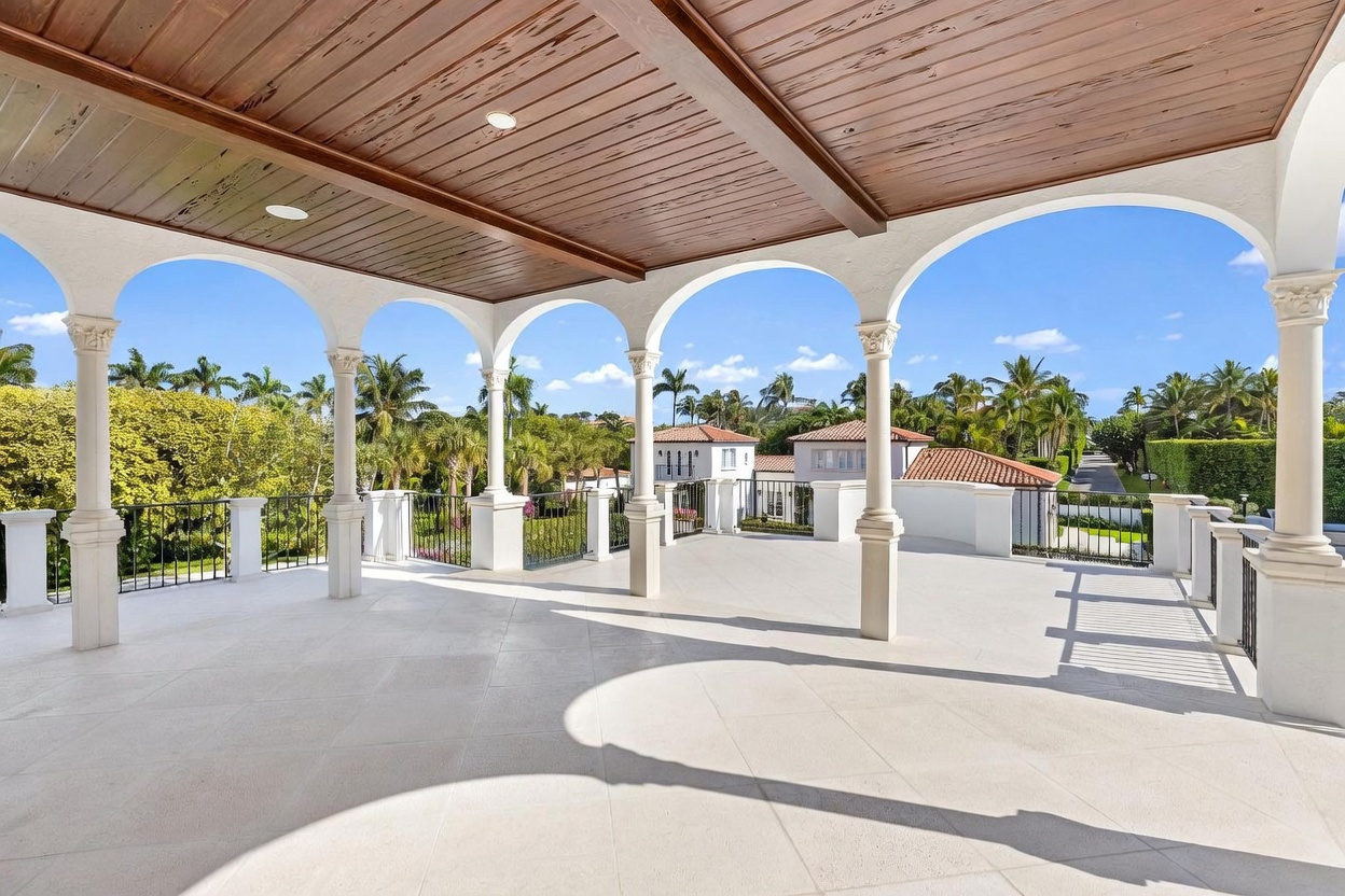 This expansive, covered balcony features a classic Mediterranean architectural style with elegant white arched columns and a warm wood-paneled ceiling. The space is finished with light-colored stone flooring and a black metal railing, offering an elevated vantage point overlooking lush tropical landscaping and neighboring properties. The open-air design creates a bright, airy atmosphere perfect for outdoor relaxation.