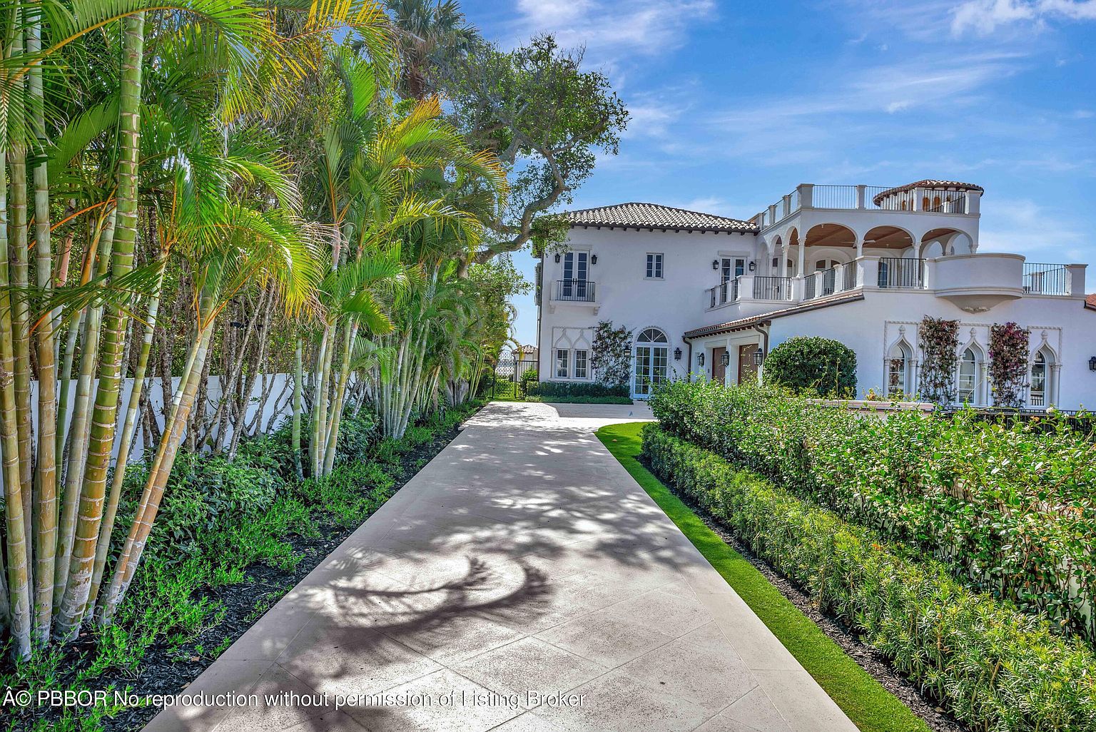 This side view of a Mediterranean-style estate showcases a long, paved driveway leading toward the rear of the property, flanked by a row of tall palm trees and a manicured hedge. The white stucco exterior features arched architectural details, multiple balconies with iron railings, and a tiled roof. The perspective captures the transition from the lush, tropical landscaping to the elegant, multi-level structure under a bright blue sky.