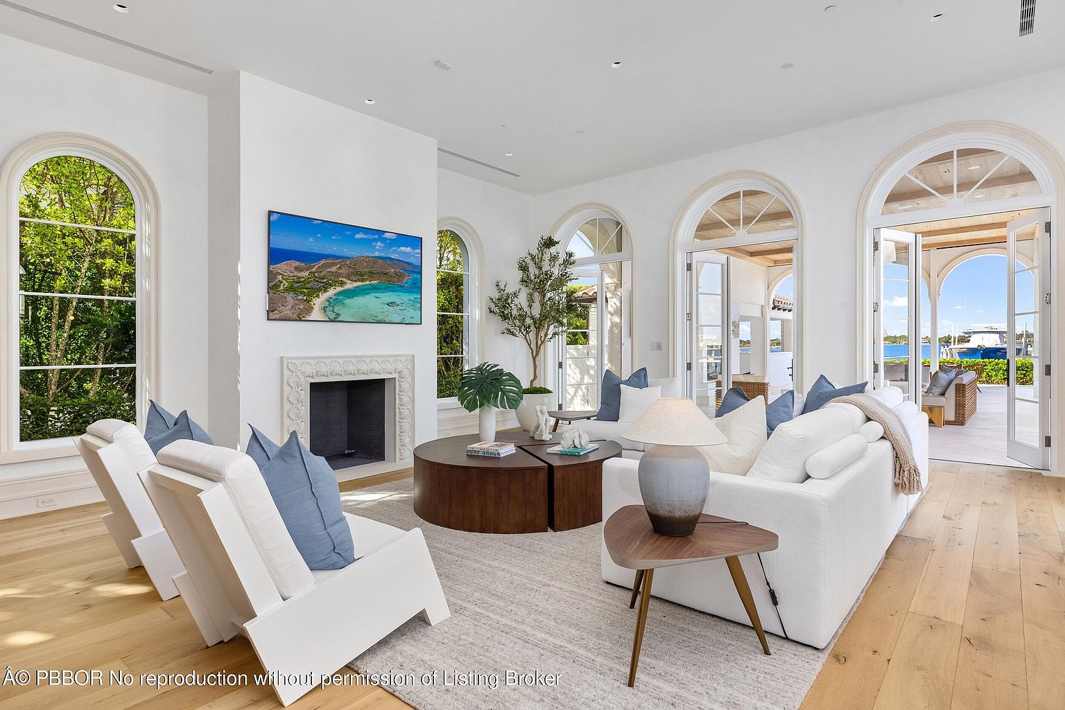 This bright and airy living room features light wood flooring and a neutral color palette, centered around a fireplace with a decorative surround and a wall-mounted television. The space is furnished with a plush white sofa and two modern armchairs, complemented by a round wooden coffee table and a textured area rug. Large arched windows and glass doors provide abundant natural light and a seamless transition to the outdoor patio area, offering a view of the water beyond.
