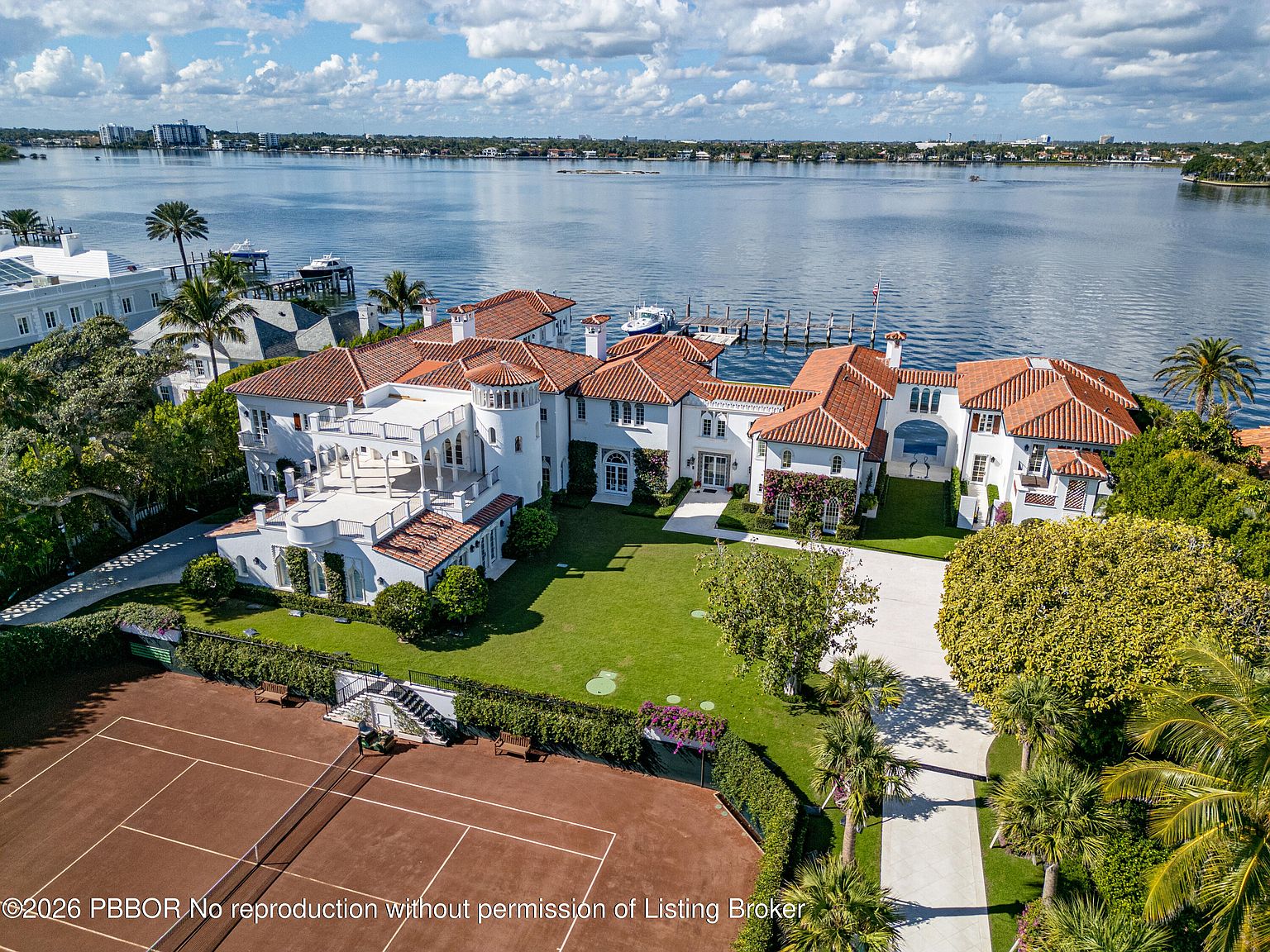 This high-angle aerial view captures a sprawling Mediterranean-style waterfront estate featuring a terracotta-tiled roof and white stucco exterior. The property includes a private tennis court in the foreground, a lush green lawn, and a deep-water dock extending into the expansive bay. The cinematic perspective highlights the home's impressive scale, its prime coastal location, and the seamless integration of luxury outdoor living spaces.