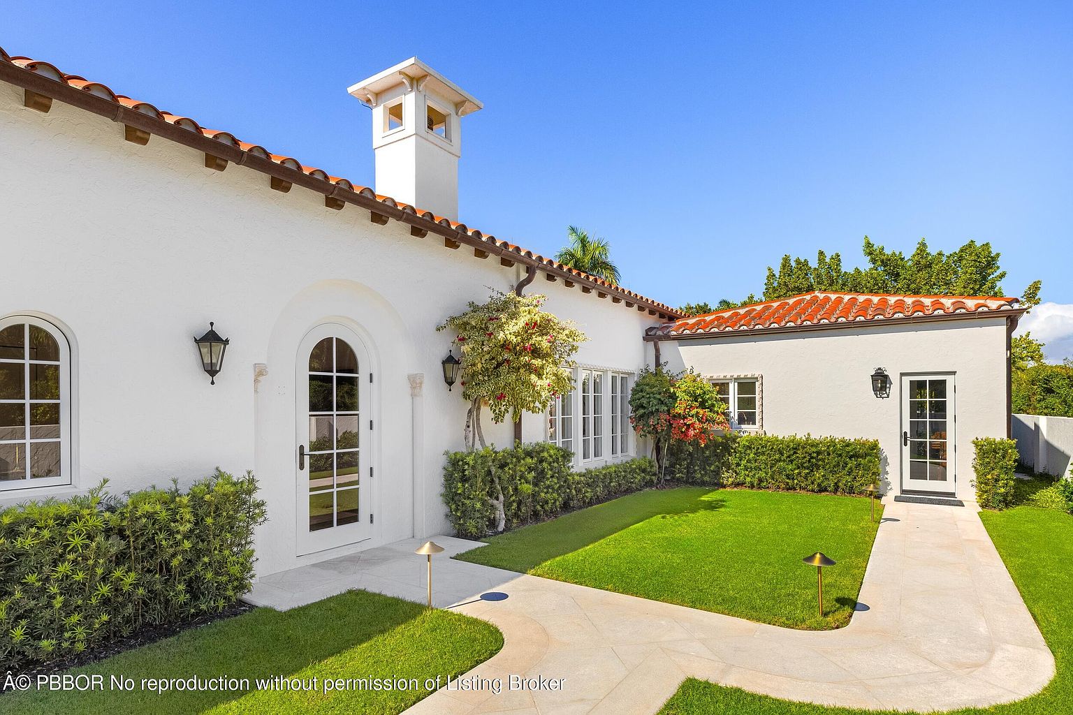 This bright, Mediterranean-style home features a crisp white stucco exterior accented by a terracotta tile roof and a prominent chimney. A stone pathway leads to a classic arched glass-paneled door, flanked by manicured hedges and a lush green lawn. The scene is captured from a low-angle perspective, emphasizing the clean architectural lines and the inviting, sun-drenched courtyard atmosphere.