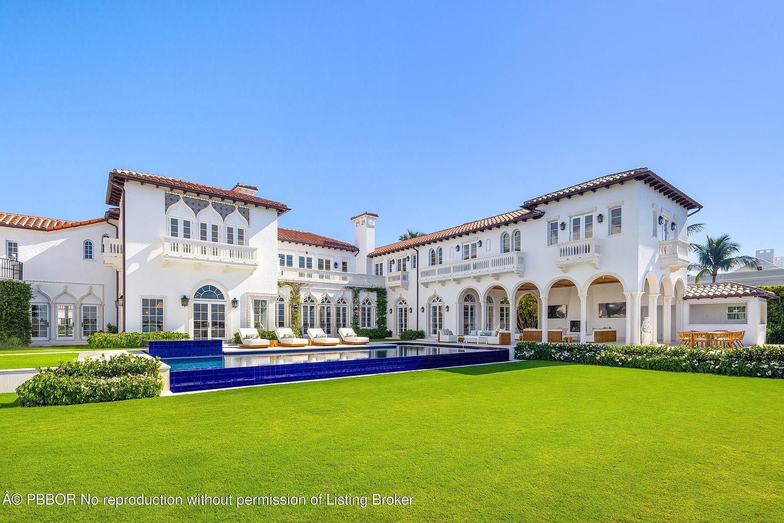 This expansive Mediterranean-style estate features a striking white stucco facade with intricate architectural detailing and a classic red-tiled roof. The rear of the home opens onto a meticulously manicured lawn and a large, rectangular swimming pool with vibrant blue tile accents. Multiple arched loggias and balconies provide seamless indoor-outdoor living spaces, creating an atmosphere of luxury and refined elegance.