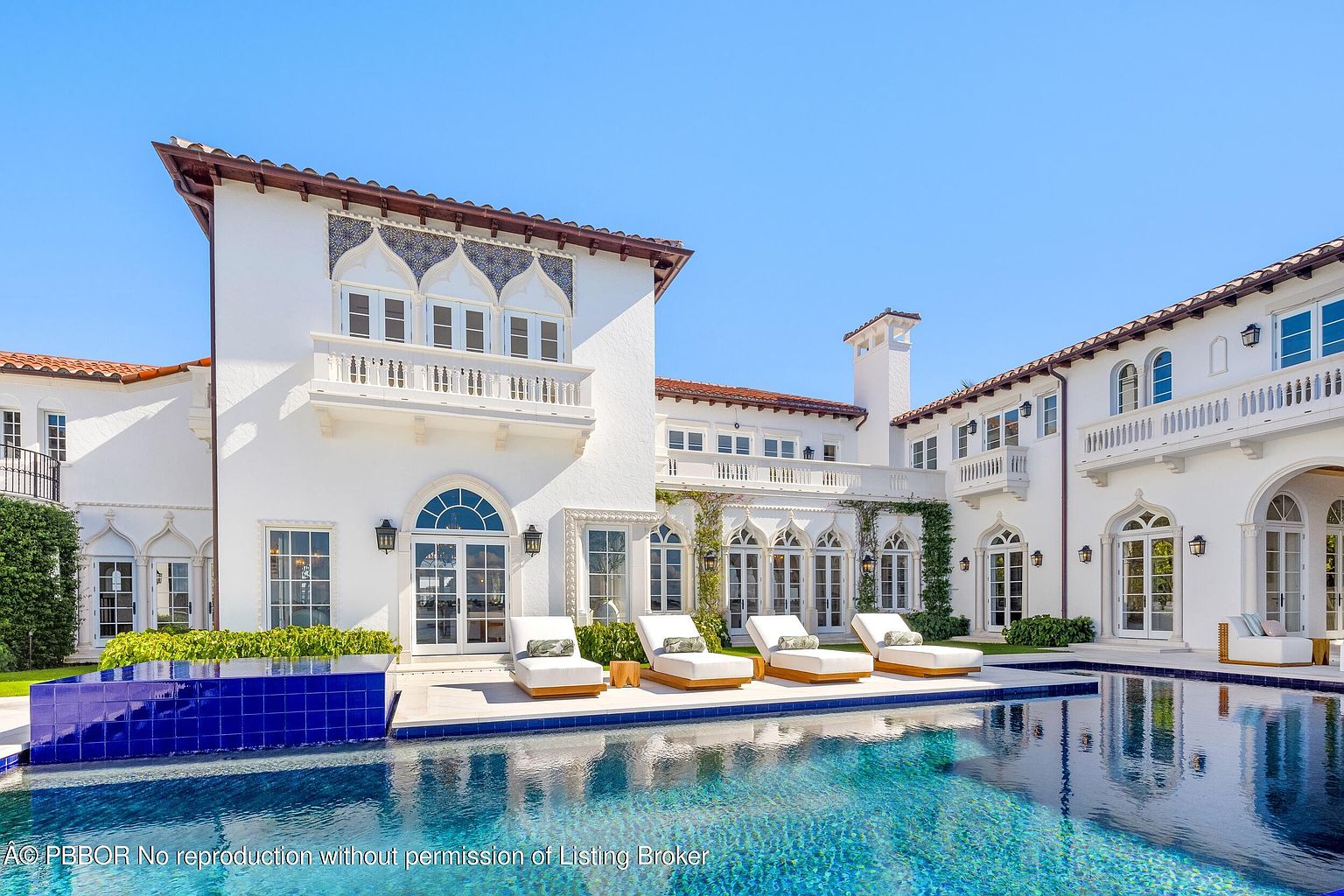 This expansive Mediterranean-style estate features a grand white stucco facade with intricate arched windows and decorative tile accents. A large, sparkling swimming pool with a raised blue-tiled water feature dominates the foreground, complemented by a row of four modern lounge chairs on the stone patio. The symmetrical architecture and lush greenery create a luxurious, resort-like atmosphere perfect for outdoor entertaining.