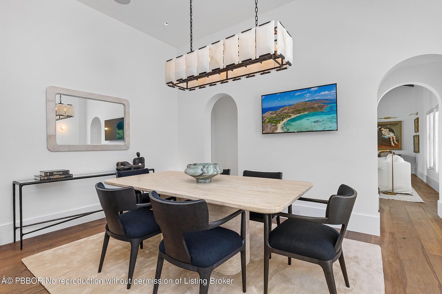 This elegant dining room features a light-toned stone table surrounded by six dark-upholstered chairs, centered beneath a large, rectangular chandelier with frosted glass panels. The room is anchored by a neutral area rug on dark wood flooring, with a sleek console table and a decorative mirror on the left wall. A television mounted on the wall displays a coastal landscape, while an arched doorway leads to an adjacent living space, creating an open and sophisticated atmosphere.