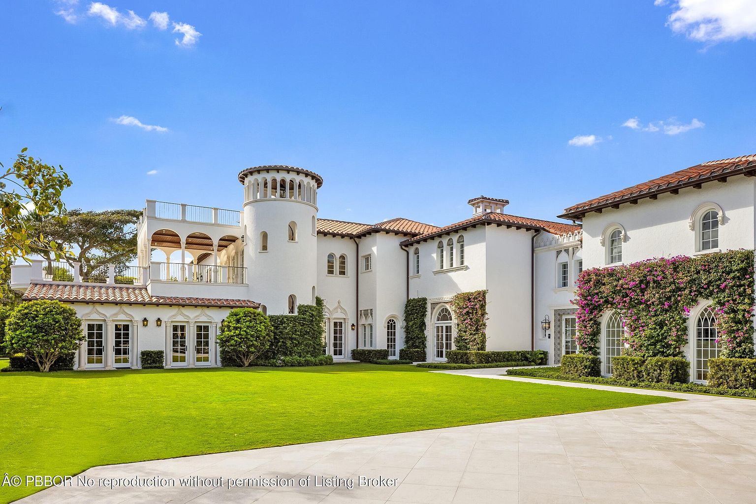 This expansive Mediterranean-style estate features a striking white stucco facade with a prominent cylindrical tower and a multi-level terrace. The property is framed by a meticulously manicured lawn and lush landscaping, including vibrant bougainvillea climbing the exterior walls. The scene is captured from a low-angle perspective, emphasizing the grand scale and architectural elegance of the residence against a clear blue sky.
