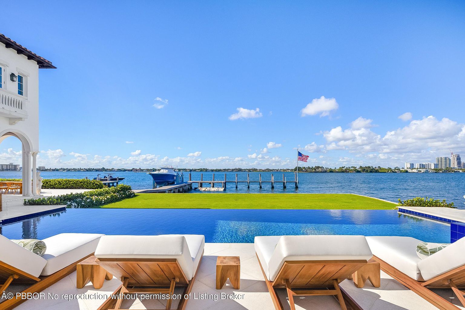 This luxurious waterfront property features a stunning infinity-edge pool that seamlessly blends into the expansive bay view. In the foreground, several wooden lounge chairs with white cushions are arranged on a stone patio, perfect for relaxing while overlooking the private dock and boat lift. The scene captures a serene, high-end coastal lifestyle with a clear blue sky and a distant cityscape on the horizon.