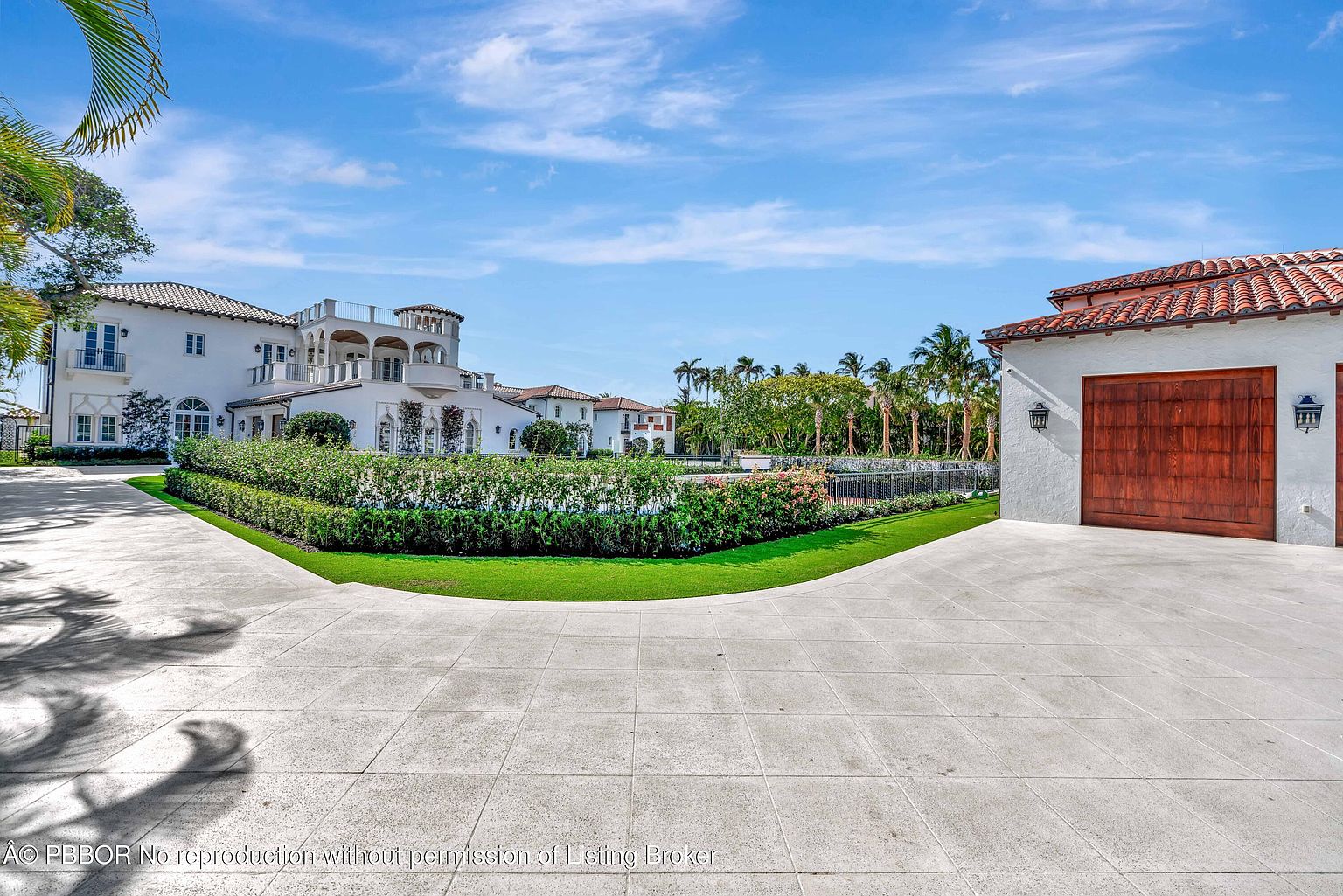 This expansive rear exterior view showcases a grand Mediterranean-style estate with white stucco walls, a terracotta tile roof, and multiple balconies. A large, paved motor court leads toward the main residence, while a manicured hedge and lawn area separate the driveway from a fenced pool area. The scene is captured under a bright, clear blue sky, emphasizing the property's luxurious scale and well-maintained grounds.