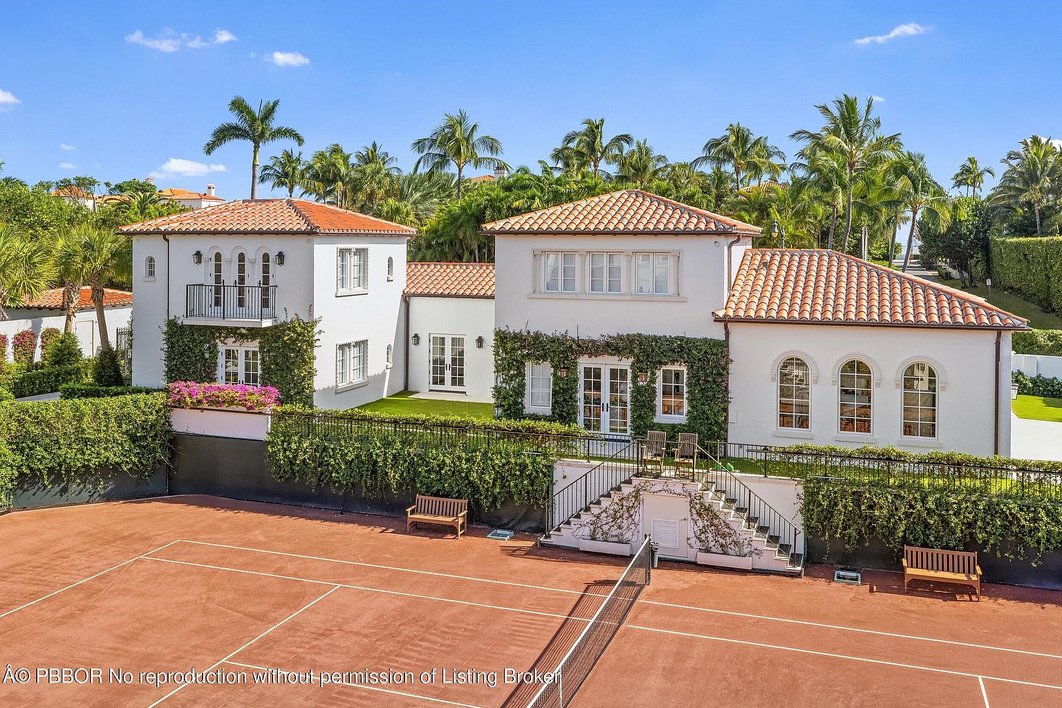 This Mediterranean-style estate features a multi-level white stucco facade with a classic terracotta tile roof, set against a backdrop of lush palm trees and a clear blue sky. The property includes a private clay tennis court in the foreground, complete with a spectator bench and a staircase leading up to the main residence. The architecture is characterized by symmetrical window placement, climbing ivy, and a second-story balcony, creating an elegant and inviting outdoor living space.