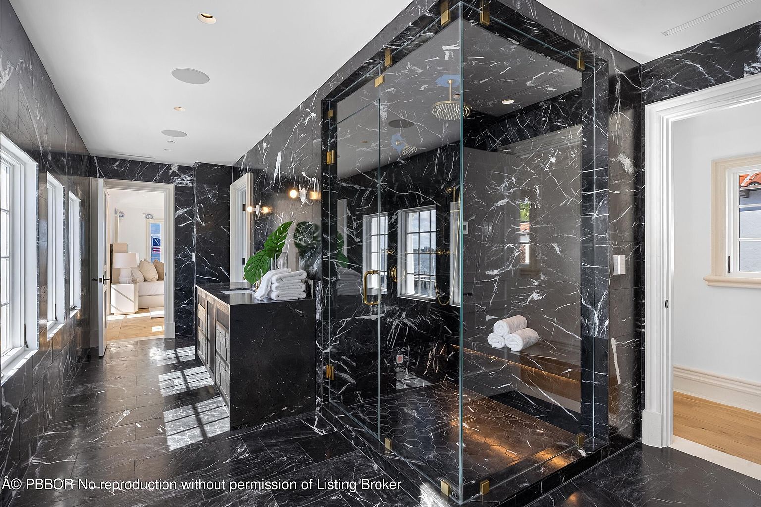 This luxurious primary bathroom is defined by its dramatic use of dark, veined stone throughout the walls and flooring. A large, glass-enclosed shower stall serves as the focal point, featuring a built-in stone bench and a rainfall showerhead. The space is brightened by natural light from multiple windows and includes a sleek vanity area, creating a sophisticated and high-end atmosphere.