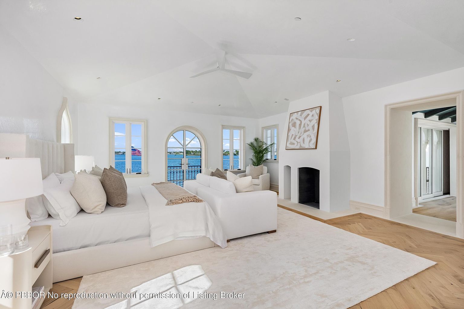 This bright and airy primary bedroom features a neutral color palette, light wood flooring, and a large area rug. The room is anchored by a plush bed and a white sofa positioned at the foot, with a tall fireplace surround serving as a focal point. Large windows and an arched glass door provide expansive water views, creating a serene and luxurious atmosphere.