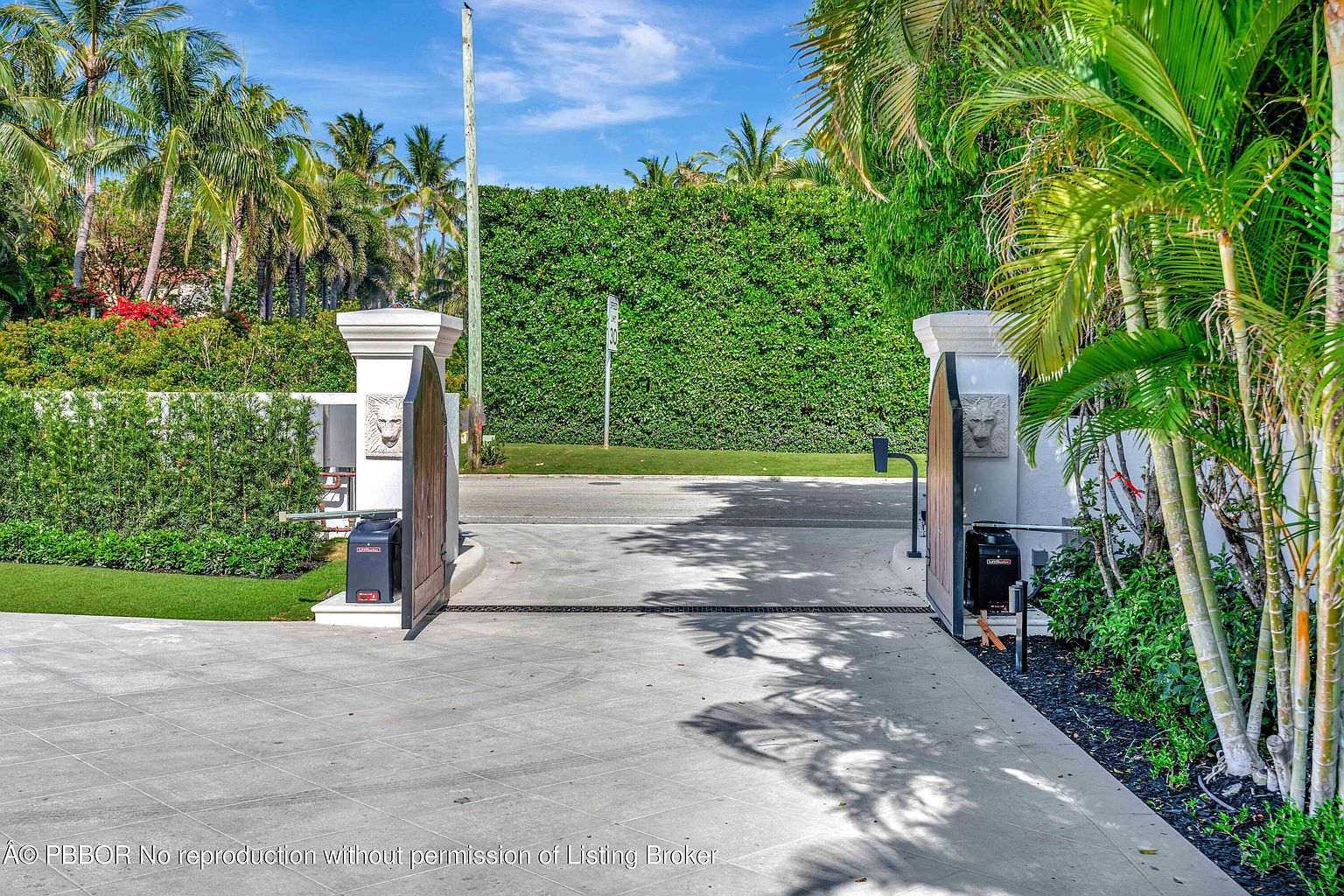 This image captures the gated entrance to a private property, featuring substantial stone pillars topped with decorative lion head accents. The driveway is paved with large, light-colored stone tiles and is flanked by lush tropical landscaping, including tall palm trees and a dense green hedge. The perspective is from inside the property looking out toward the street, emphasizing the privacy and security of the gated access.