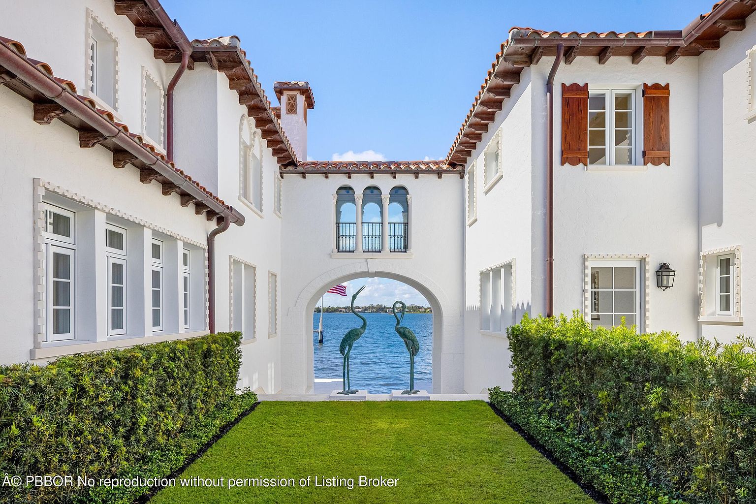 This Mediterranean-style estate features a symmetrical white stucco facade with a central arched breezeway that frames a picturesque view of the water. A manicured lawn leads toward the arch, which is flanked by two bronze crane sculptures, while the structure is accented by dark wood shutters and terracotta roof tiles. The composition creates a grand, open-air corridor that seamlessly connects the residence to the waterfront.