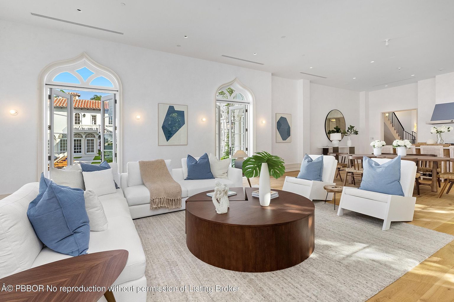 This bright and airy living room features a neutral color palette with white sofas, light wood flooring, and a large circular wooden coffee table as a central focal point. The space is accented by blue throw pillows and abstract artwork, while tall arched glass doors provide a seamless transition to the outdoor patio area. The open-concept layout flows naturally into a dining area in the background, creating a sophisticated and inviting atmosphere.