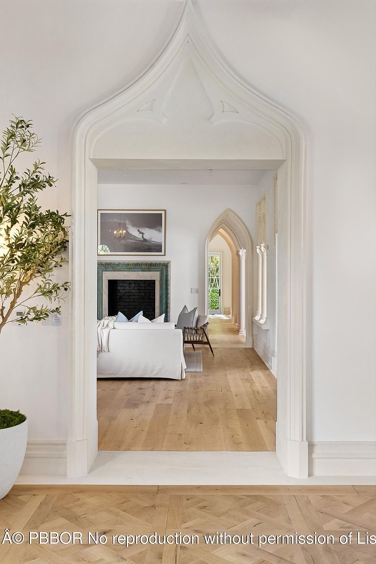This perspective captures a view through a striking, pointed gothic-style arched doorway into a bright living area. The foreground features light-toned wood flooring in a herringbone pattern, while the background reveals a cozy seating area with a fireplace and a secondary arched hallway leading to an exterior door. The space is characterized by clean white walls, elegant architectural detailing, and a sophisticated, airy aesthetic.