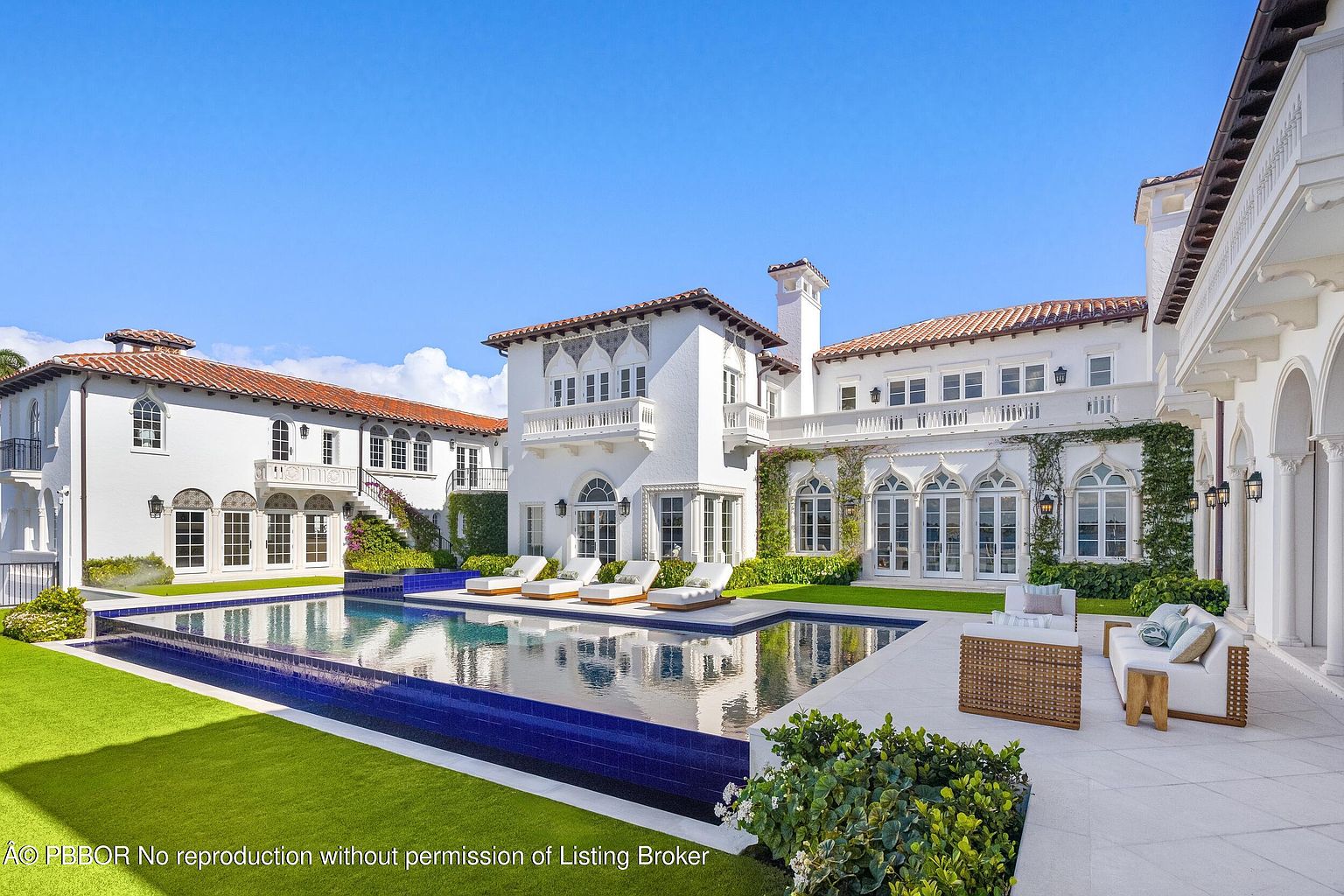 This expansive Mediterranean-style estate features a stunning rear view showcasing a large, rectangular swimming pool with a deep blue tiled edge. The white stucco architecture is complemented by arched windows, terracotta roof tiles, and lush green landscaping, creating a luxurious and serene atmosphere. A spacious stone patio surrounds the pool, furnished with lounge chairs and outdoor seating, perfect for entertaining in this bright, sun-drenched setting.