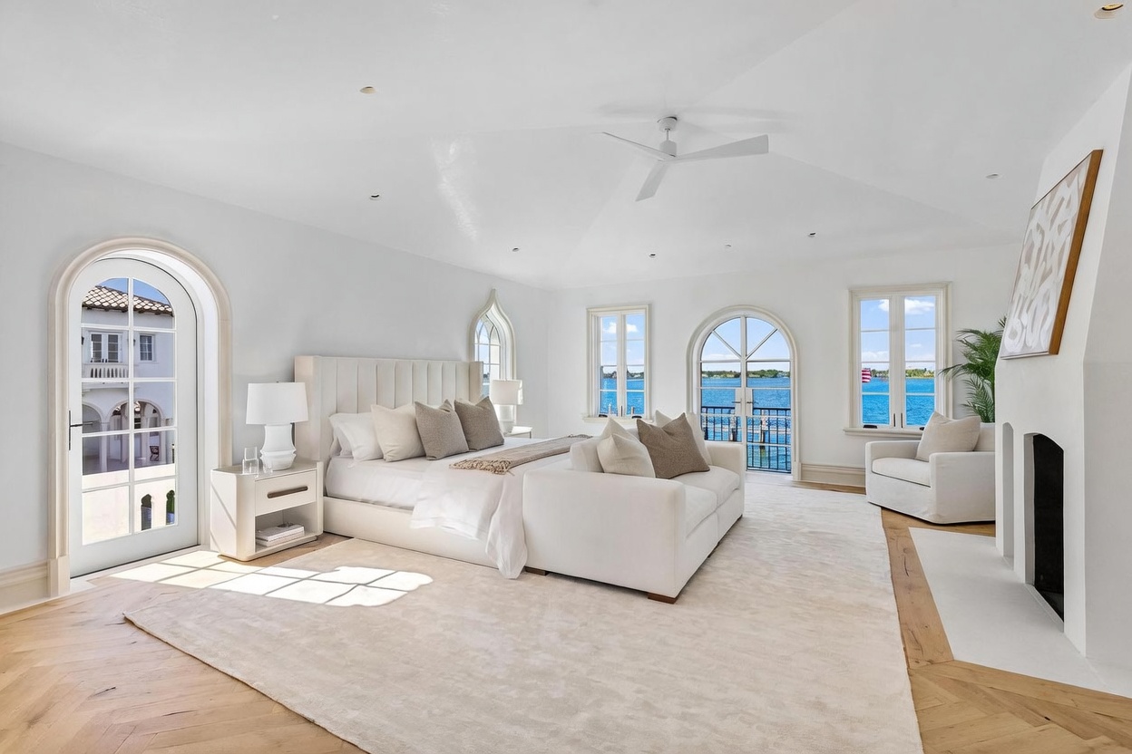 This expansive primary bedroom features a bright, airy aesthetic with white walls, light wood flooring, and a vaulted ceiling. The room is anchored by a large upholstered bed with a matching sofa at its foot, positioned to face a fireplace with a tall, minimalist surround. Multiple arched windows and a glass door provide abundant natural light and panoramic views of the water, creating a serene and luxurious atmosphere.