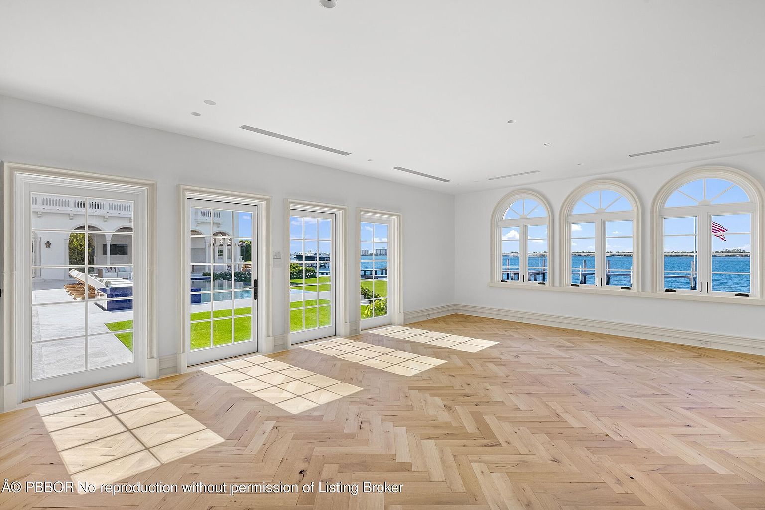 This bright, expansive living space features light wood flooring laid in a classic herringbone pattern and crisp white walls. The room is defined by multiple glass doors and large arched windows that provide abundant natural light and panoramic views of the water and pool area. The minimalist design emphasizes the architectural connection between the interior living space and the outdoor waterfront environment.