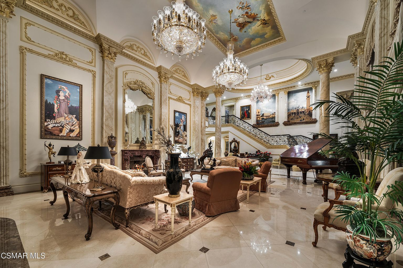 This opulent, grand living room features soaring ceilings with a detailed fresco, multiple crystal chandeliers, and polished marble flooring. The space is furnished with ornate, traditional-style seating, a grand piano, and a fireplace, all framed by classical columns and large-scale artwork. The perspective is a wide-angle shot from the entryway, capturing the immense scale and luxurious, palatial aesthetic of the home.
