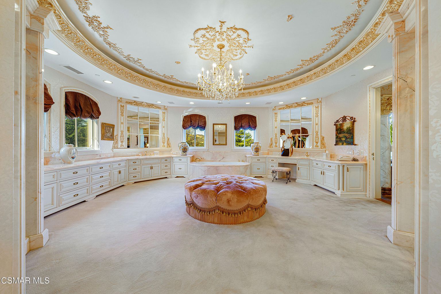 This opulent primary bathroom features a grand circular layout with a central tufted velvet ottoman and a luxurious soaking tub nestled against the back wall. The space is adorned with intricate gold-leaf ceiling molding, a crystal chandelier, and custom white cabinetry with marble countertops, creating an atmosphere of classic elegance and high-end sophistication. The perspective is wide and centered, showcasing the symmetrical design and the expansive, carpeted floor plan.